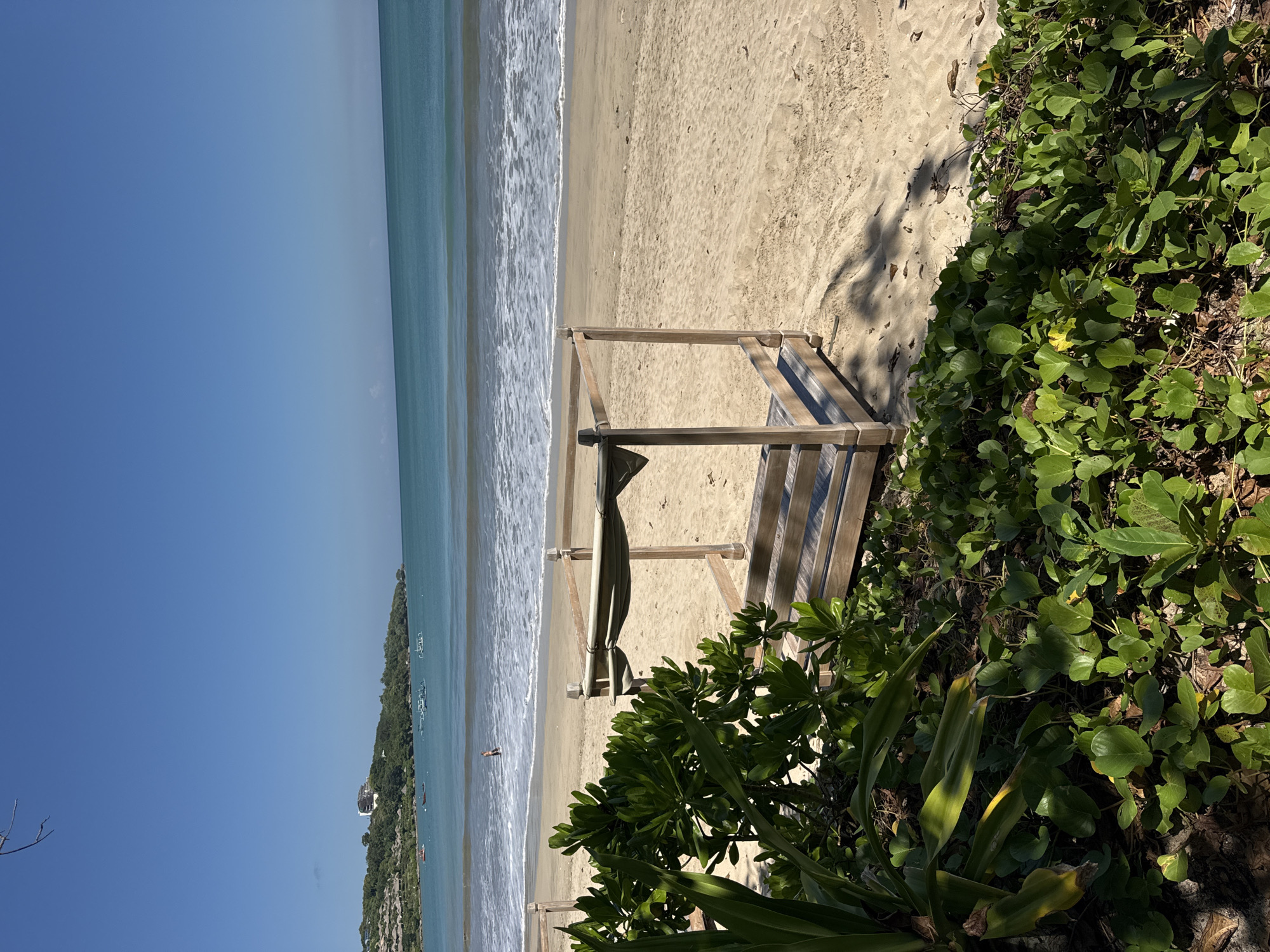 The image shows a beach scene in Kuta Selatan, Indonesia. The beach consists of golden sand bordered by turquoise water with gentle waves. A wooden structure, possibly a beach cabana frame, stands partially on the sand and partially amidst lush, green vegetation at the edge of the beach. A small coastal hill covered in trees is visible in the distance. The sky is clear and blue. A single person is present in the water.