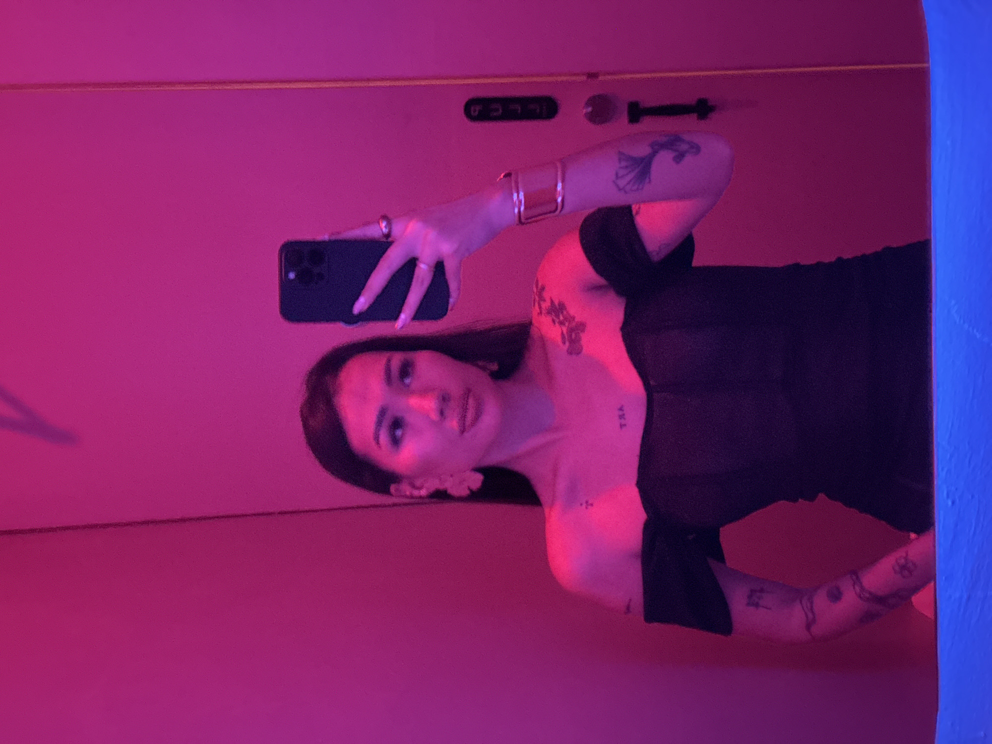 A woman with dark hair is taking a selfie in a room lit with pink and blue lights. She is wearing a black dress with short sleeves and has tattoos on her arms, shoulder, and chest. She is holding a black smartphone in her left hand and looking at the screen. The room appears to be a hotel room, possibly in North Kuta, Indonesia.