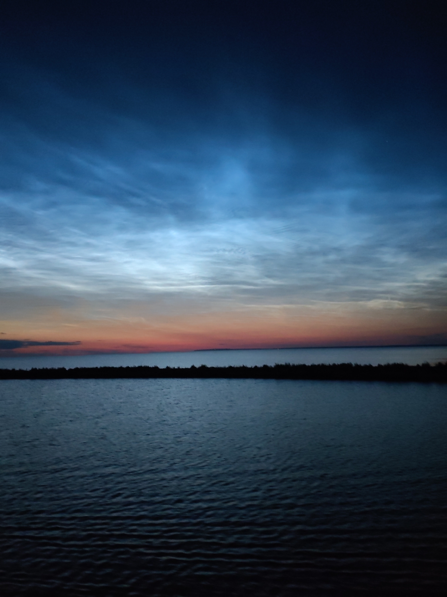 The image depicts a serene natural scene at dusk over the waters of Ozernoe Ust'e, Russia. The sky is a gradient of deep blue at the top, transitioning to lighter blues and then a band of orange and pink hues near the horizon, indicative of a sunset or twilight. Wispy, bright clouds, possibly noctilucent clouds, are visible in the upper portion of the sky, catching the last light. A dark, silhouetted landmass or shoreline runs horizontally across the middle of the image, separating the sky from the body of water in the foreground. The water itself is dark and reflects the dim light of the sky, with subtle ripples on its surface. The overall atmosphere is calm and tranquil, with no people or artificial structures clearly visible.