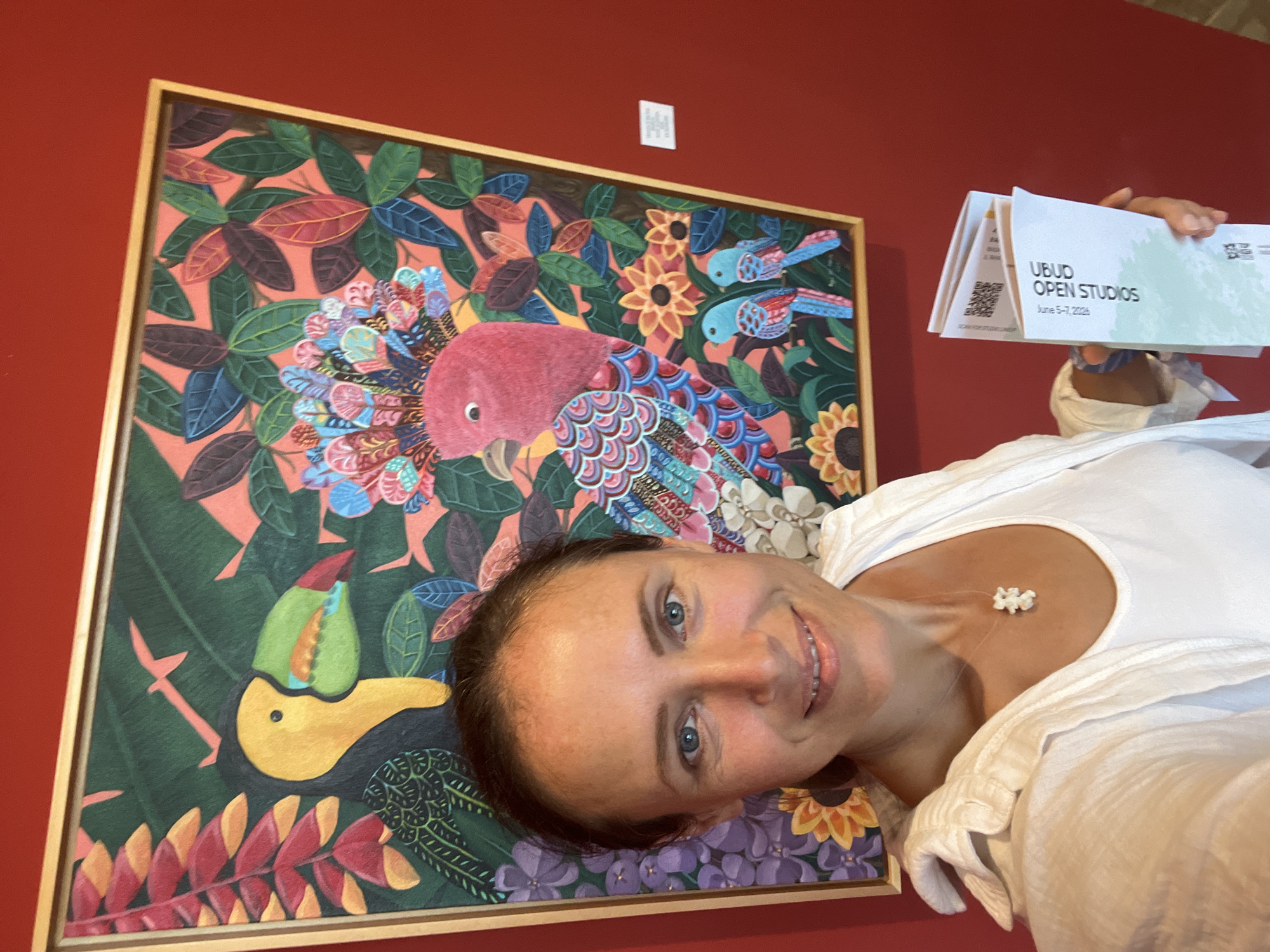 A light-skinned adult female is captured in a selfie, positioned centrally in front of a vibrant, framed painting of tropical birds and foliage, against a deep red wall. She has dark hair pulled back, blue eyes, and a pleasant smile. She is wearing a white sleeveless top under an open white textured long-sleeved shirt. A delicate white pendant is visible on a thin necklace around her neck. In her left hand, she holds a folded white brochure displaying "UBUD OPEN STUDIOS" and "June 3-5 2023," indicating the location is in the city of Ubud District, Indonesia.
The painting, set in a narrow gold-toned frame, depicts a vivid tropical scene. On the left, a large yellow toucan with a green and red beak is shown amidst green leaves and red hanging flowers. On the right, a prominent pink parrot with an elaborate, multi-colored feathery crest and patterned body (featuring shades of blue, pink, and red) is surrounded by broad green and dark red leaves and yellow sunflower-like blossoms. Two smaller blue and red birds are perched below the parrot. The background foliage ranges from dark green to lighter pink-orange hues. The scene takes place indoors, characterized by a uniform deep red wall. A small, unreadable white label is affixed to the wall to the right of the painting.