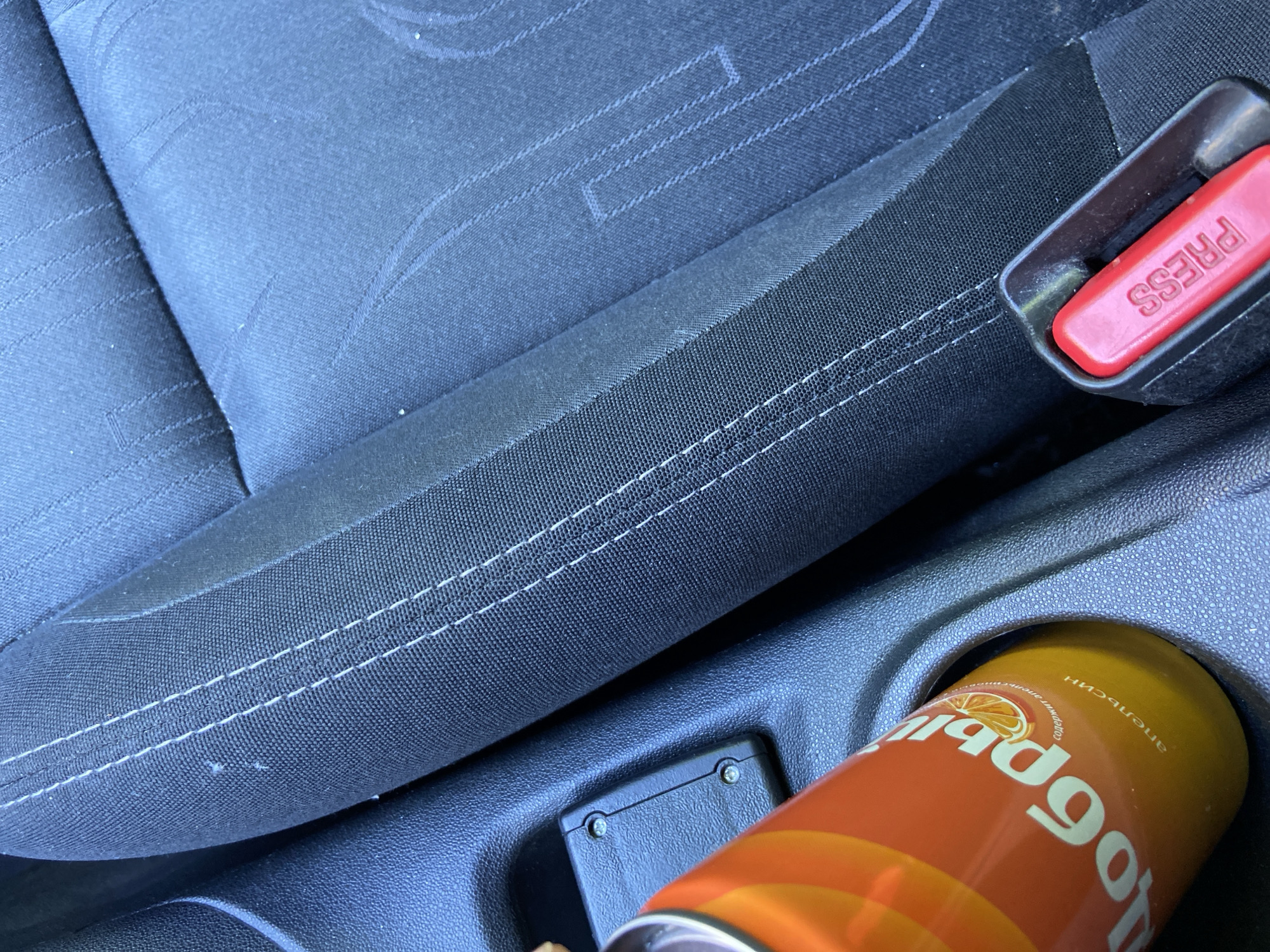 The image shows the interior of a vehicle, likely a car, with a focus on the central console area and part of a car seat. The car seat is upholstered in dark fabric with decorative stitching. A bright orange soda can with Russian text, reading "Добры" (Dobry) and "апельсин" (apelsin - orange), is placed in a cup holder on the central console. To the right of the cup holder is a black seatbelt buckle with a red "PRESS" button. The background consists of the car's interior, including plastic trim and the car seat. There are no people visible in the image. The scene suggests a personal, everyday setting within a vehicle. The reference to Saint Petersburg, Russia, is not directly evident from the visual information in the image itself.