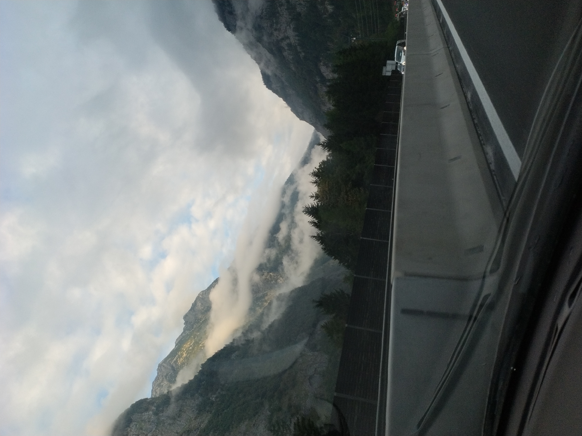 The image captures a scenic view of a mountain pass from the perspective of a vehicle on a highway. The road in the foreground features a concrete barrier and lane markings. Beyond the barrier, a dense forest of evergreen trees lines the base of imposing, mist-shrouded mountains. The sky is overcast with scattered clouds. A few vehicles are visible on the road ahead, including a white van. The overall impression is one of natural grandeur and a journey through a mountainous region. The location is identified as Wimm, Austria, likely on a highway traversing the Alps.