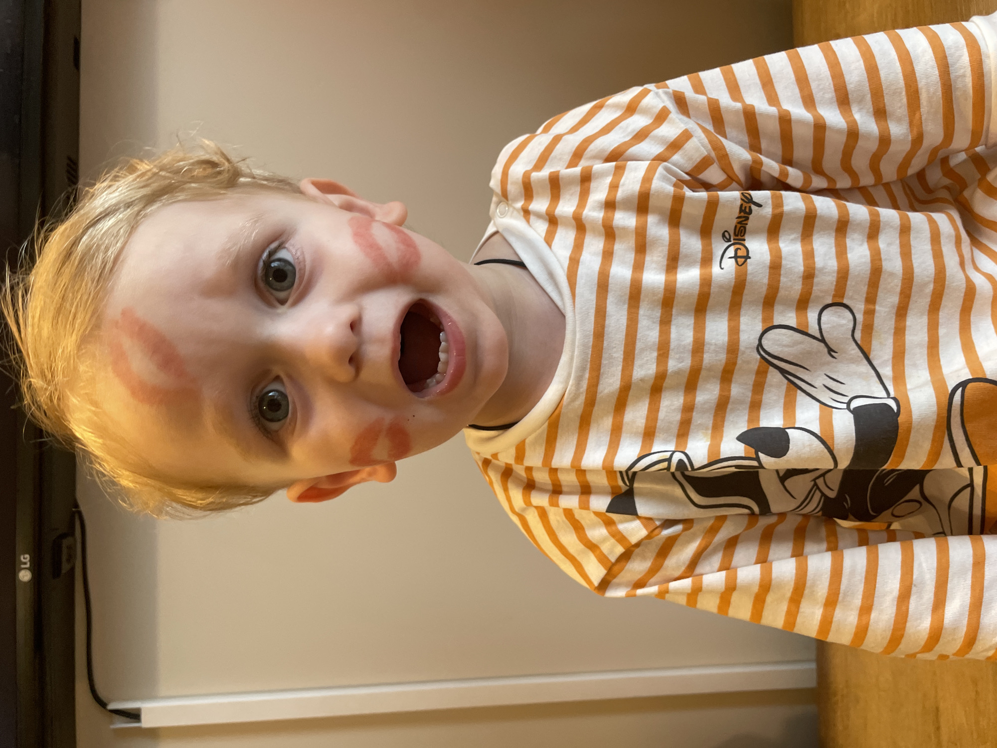 A blond child with blue eyes is looking directly at the camera, with an open mouth expression, showing his teeth. He has lipstick marks on his left cheek and forehead. He is wearing a white and orange striped long-sleeve shirt with a Disney Mickey Mouse design. The background shows a white wall, a dark-colored LG monitor, and a brown wooden surface. The location is unknown.