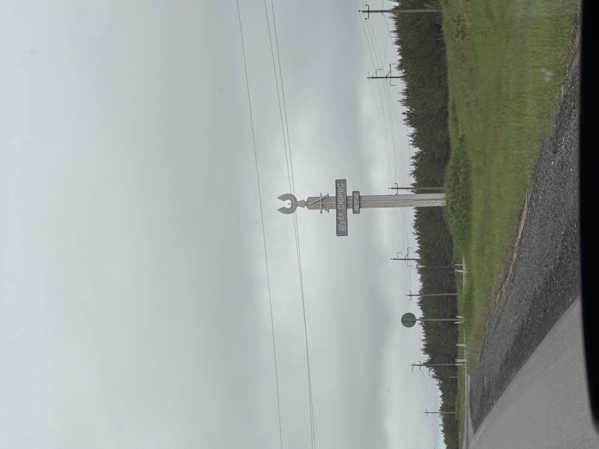 A roadside scene captured on an overcast day in Alnashi, Russia, shows a unique monument next to a paved road with a gravel shoulder. The sky is uniformly grey, indicating cloudy weather.
The primary subject is a tall, light-colored monument or sign. At its apex, there is a large, metallic crescent wrench shape. Below this, the monument features several rectangular panels. The most prominent visible text on one of these panels is "ШУӞ ӦРЧЕС" (Shuzh Orches), written in Cyrillic script. Below this, another panel repeats "ШУӞ" (Shuzh), with further text below it that is partially obscured and difficult to read due to blur and angle. This text is in the Udmurt language, consistent with the Alnashi location in the Udmurt Republic.
To the right of the monument, extending into the distance, is a row of power lines supported by wooden poles. A dense line of green trees or shrubs forms a natural border behind the road and monument. Further down the road, another circular road sign is visible, though its specific markings are indistinguishable. The foreground shows green grass leading to the treeline. No people or active events are depicted, presenting a static landscape view.