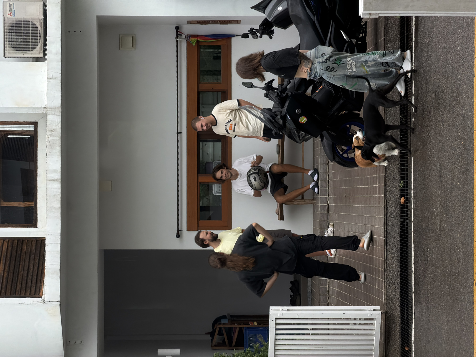 The image depicts an outdoor scene in front of a building, likely residential, in Chang Phueak, Thailand. A group of four young adults and two dogs are present. In the foreground, a black motorcycle is parked, and to its right, a young woman wearing a black crop top and patterned jeans stands with her back to the camera. Two dogs, one small and light-colored, and a larger black dog, are interacting near the motorcycle and the woman.
Behind the motorcycle, two young men are standing, one in a yellow t-shirt and shorts, and the other in a white t-shirt and shorts, holding a white helmet. A third young man, also in a white t-shirt and shorts, is seated on a wooden bench in front of a window. Another young woman, with long brown hair, is standing on the left side of the image, facing away from the camera, wearing a black t-shirt and dark pants.
The building features a white facade with wooden window frames and louvers. An outdoor air conditioning unit is visible on the upper part of the building. The ground is paved with brick-like tiles, and a metal drain runs along the edge of the paved area, bordering a wet asphalt road with a yellow line. To the far left, a white metal gate and a shelving unit with various items are visible. The overall atmosphere appears casual, possibly a gathering or a break.