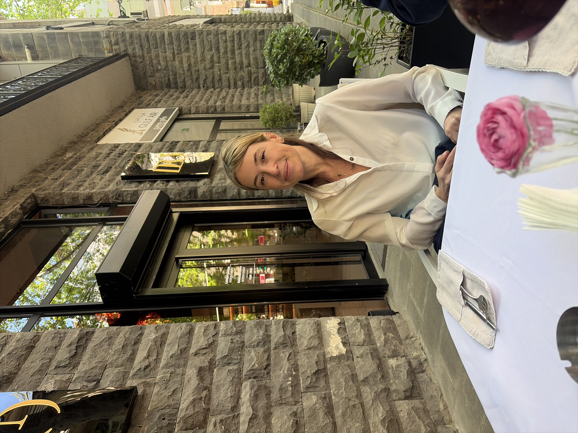 A woman is sitting at a table outside the restaurant "Gulf Air" in Tbilisi, Georgia. The restaurant is a stone building with large windows. The woman is wearing a white shirt. On the table in front of her are a napkin, silverware, a pink flower in a vase, and a glass of wine.