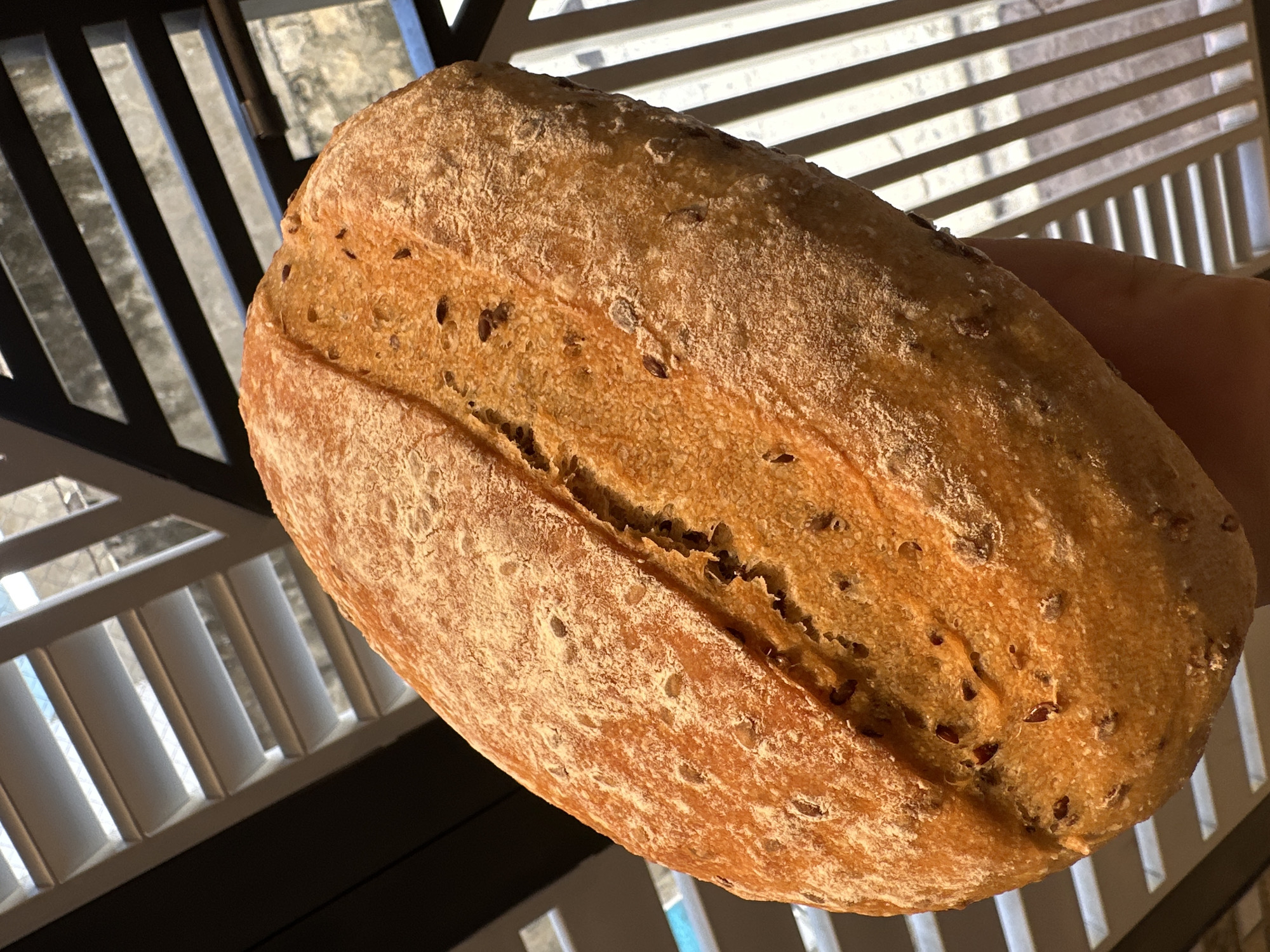 A close-up shot shows a loaf of bread with visible seeds in its crust. The bread appears golden brown and has a cracked surface. The background is composed of a partially visible window blind with alternating light and dark stripes. The bread is held by a person, with a part of the hand visible. The environment does not indicate a specific location in Bac Nha Trang Ward, Vietnam. No specific actions or interactions are taking place.