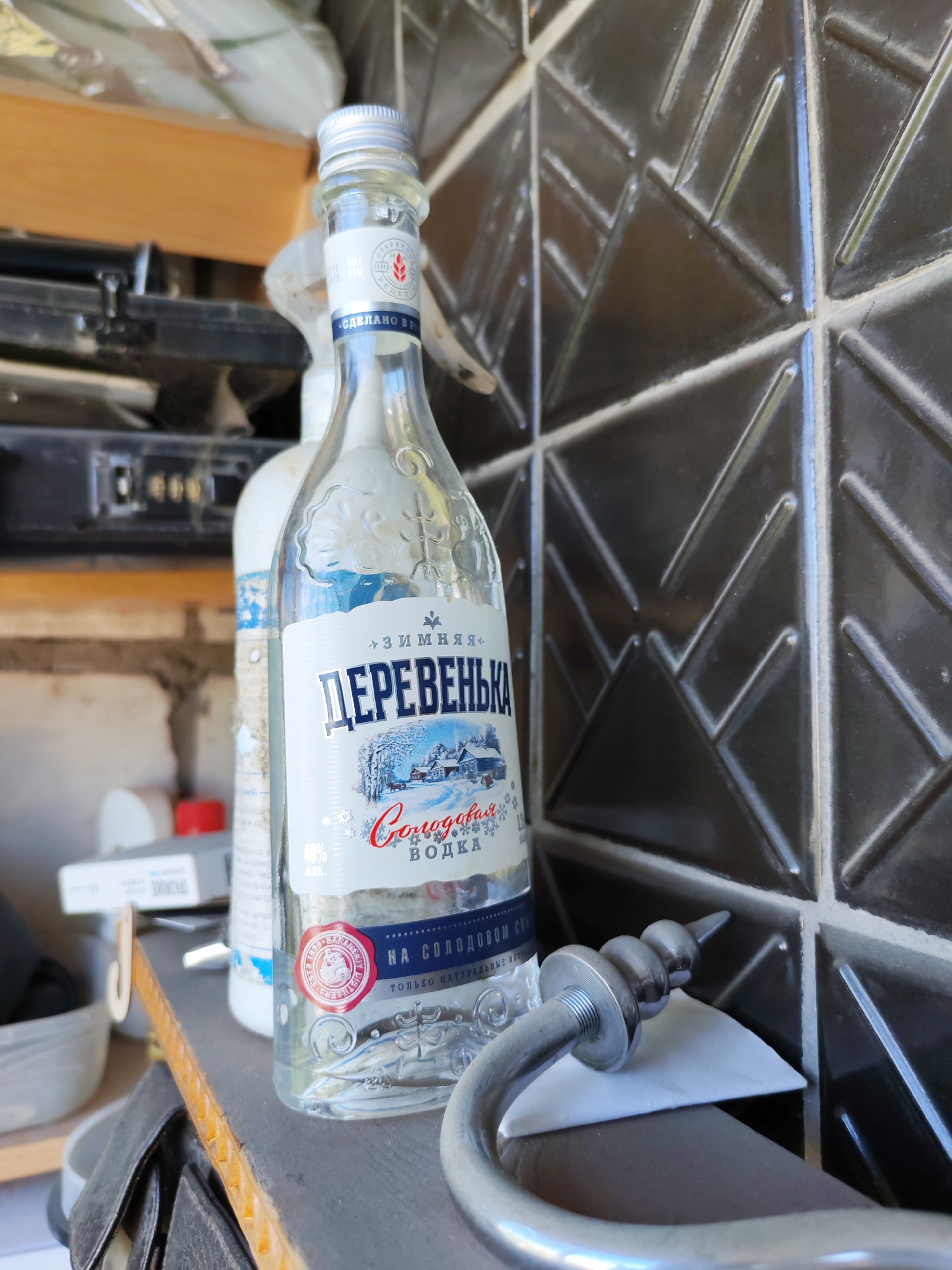 A clear glass bottle of vodka, labeled "Деревенька" (Derevenka) in Russian Cyrillic with a winter village scene on its label, stands upright on a shelf. The bottle is positioned in the foreground, slightly to the left of center, against a dark, patterned tiled wall. To the left of the vodka bottle, partially visible, is another bottle, also white, with a blue label. The shelf itself appears to be made of wood and is worn. Behind the bottles, a shelf with various objects, possibly electronics or stored items, is visible. To the right of the vodka bottle, a curved metal object, resembling a handle or part of a fixture, rests on a white piece of paper. The overall environment suggests an indoor setting, likely a pantry or storage area, and no specific indicators point to the city of Novosibirsk, Russia.