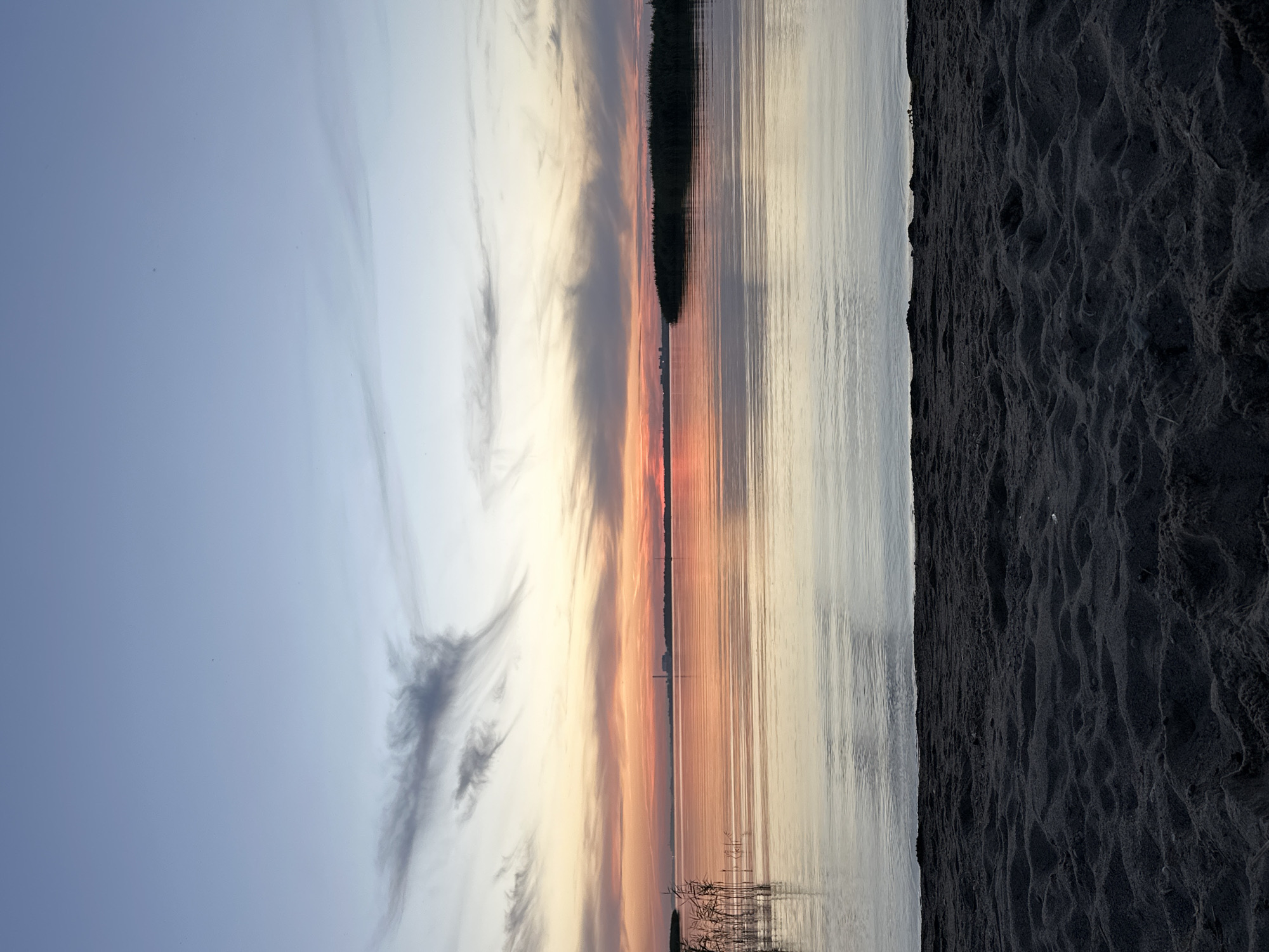 A sunset paints the sky in hues of orange and pink over a calm body of water. The foreground consists of dark, textured sand. In the distance, a silhouetted treeline forms the horizon, with a few distant buildings visible. Wispy clouds stretch across the sky, reflecting the sunset colors on the water's surface. Reeds grow along the left bank of the water.