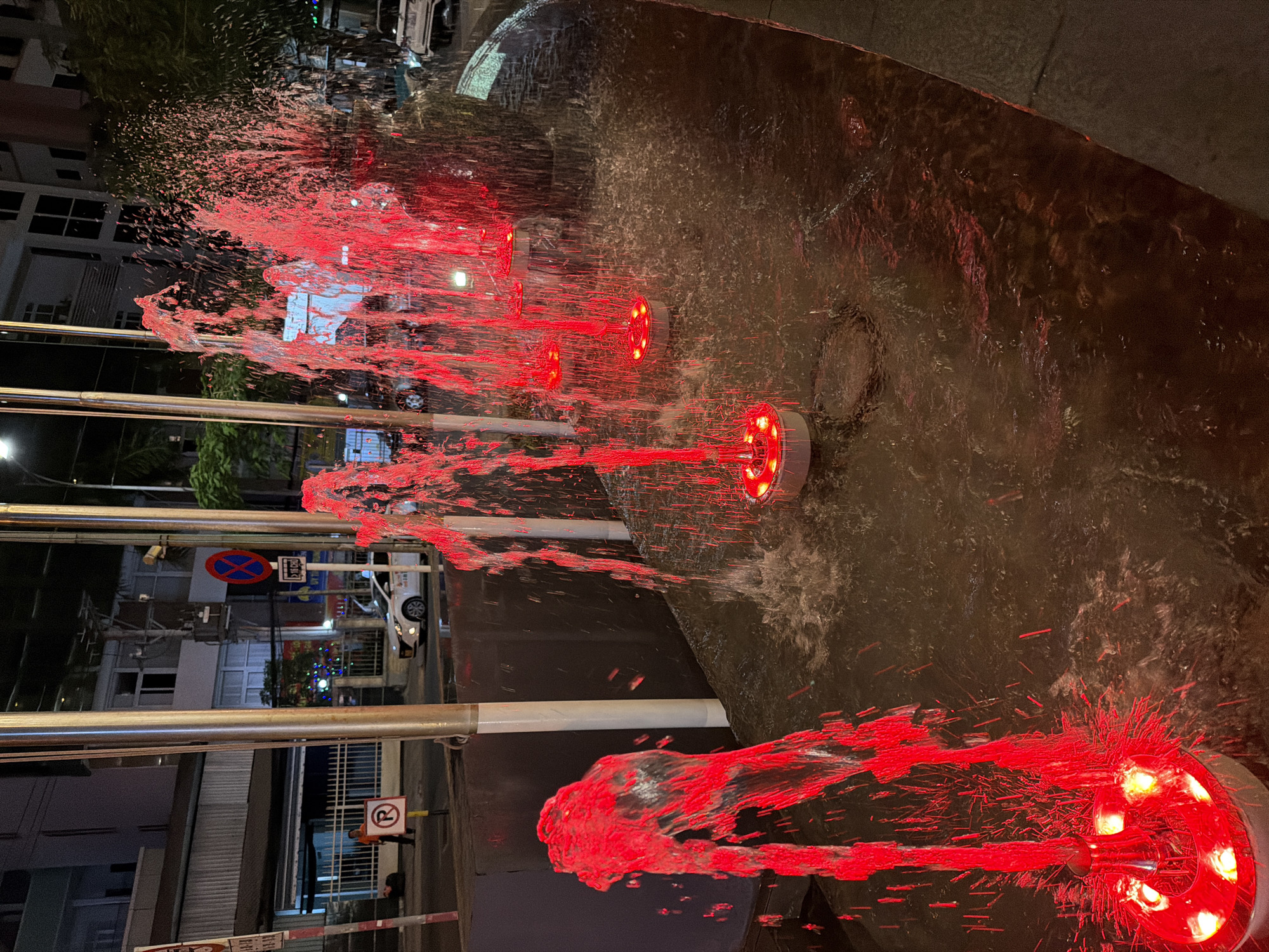 The image shows a fountain with red lights at night in Nha Trang Ward, Vietnam. The fountain has multiple spouts of water, each illuminated by a red LED light, creating a dramatic and vibrant effect. The water is spraying outwards, with droplets catching the light and scattering, forming a dynamic display.
In the background, there are buildings with illuminated windows and some greenery. Street signs are visible, including a "No Parking" sign with a blue circle and a red diagonal line, and another sign with a "R" symbol indicating a roundabout. The presence of streetlights and the overall dark sky suggest it is nighttime. The setting appears to be a public area, possibly a plaza or a street, given the presence of road signs and vehicles in the background. The overall mood is lively and modern, with the artificial lighting creating a festive atmosphere. There are no people clearly visible in the frame, focusing the attention on the water and light spectacle.