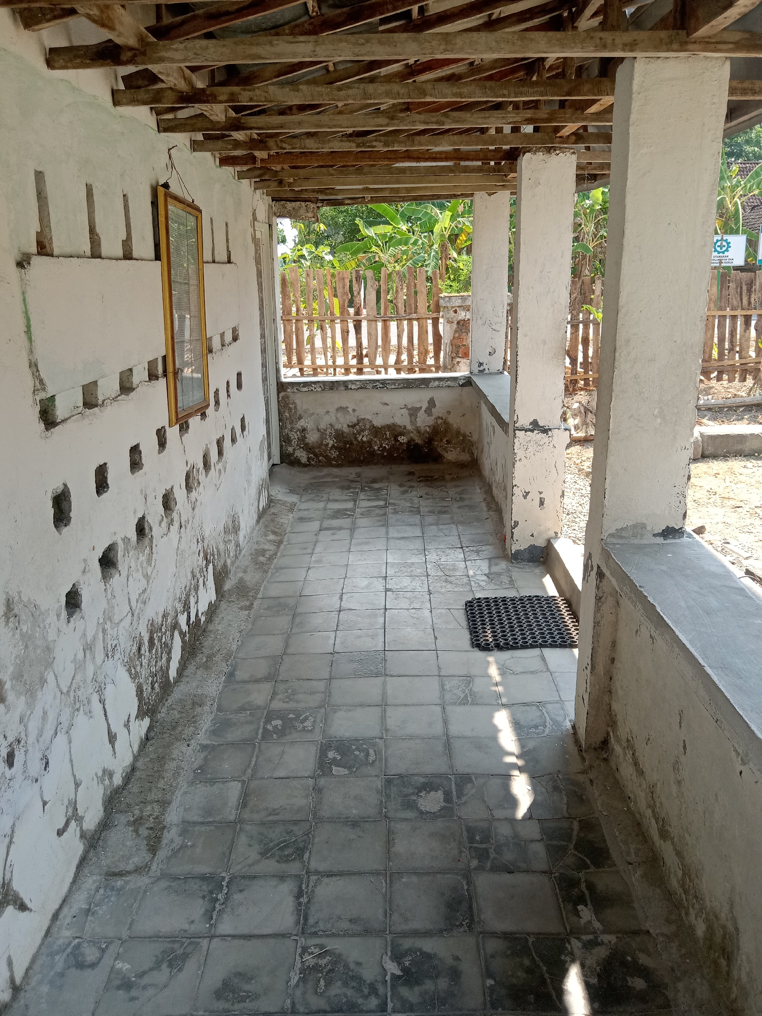 The image depicts an outdoor corridor with a tiled floor. The corridor is partially covered by a wooden roof supported by concrete pillars. The walls are white and appear weathered, with several small square openings visible on the left wall. A framed picture hangs on the left wall. A black doormat is placed on the floor near the pillars. In the background, a wooden fence encloses an area with vegetation, including banana trees, and a sign with text and a logo is visible on the right. The scene appears to be in an outdoor setting, possibly a rural area or the outskirts of a city.