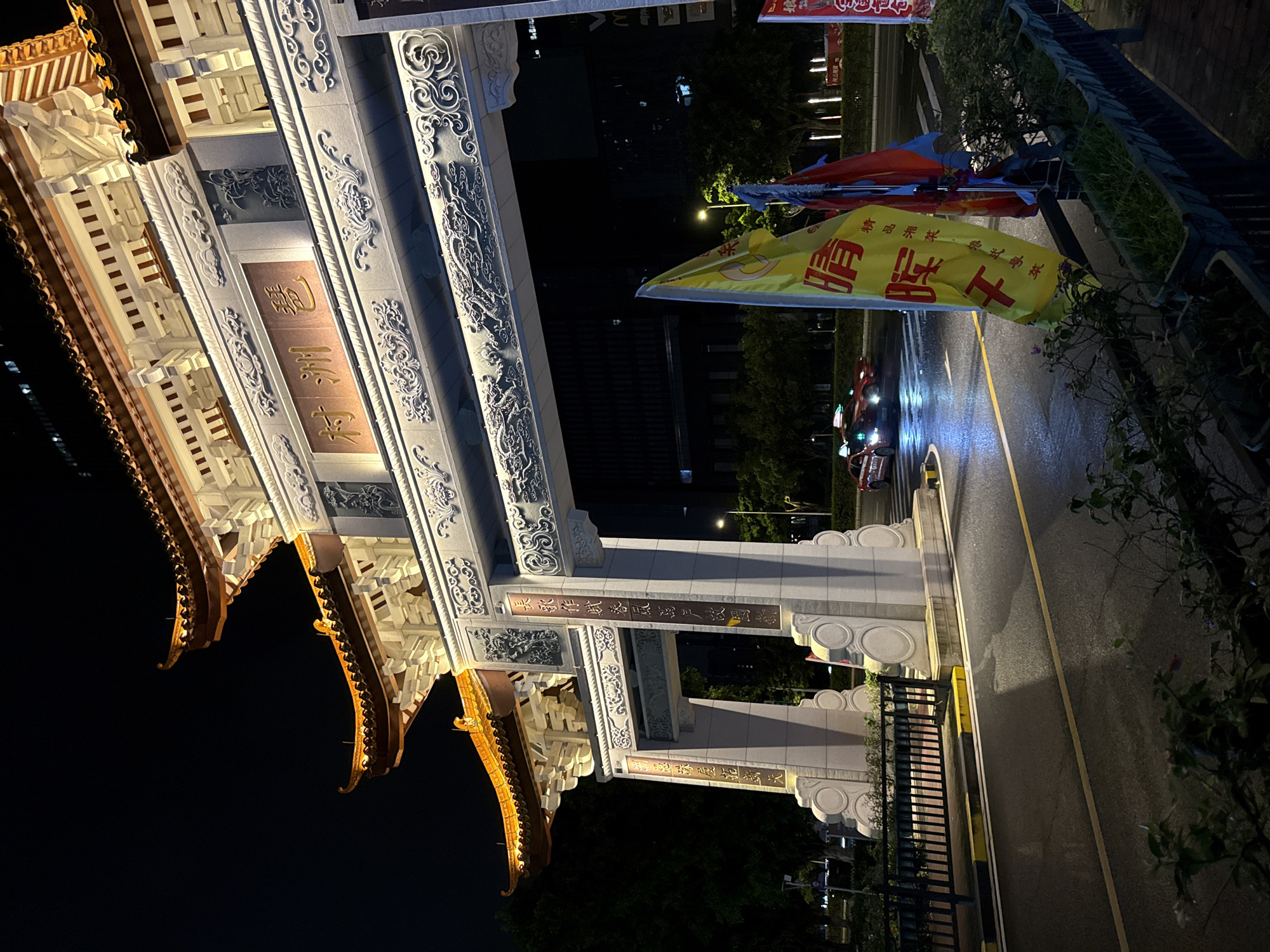This is a nighttime, medium-shot photograph of a large, ornate archway in Haizhu District, China. The archway is constructed of stone and features traditional Chinese architectural elements, including multiple tiered roofs with upturned eaves illuminated by warm orange light. Intricate carvings of dragons and swirling patterns adorn the archway's structure. Chinese characters are visible on a golden plaque above the main passageway, and vertical calligraphy is inscribed on the supporting pillars.
In the background, a dark, modern building stands tall, its windows dimly lit. The street in front of the archway is wet, suggesting recent rain, with reflections visible on the asphalt. A yellow taxi with its illuminated roof sign is driving through the archway. Several flags, including a prominent yellow one with red Chinese characters and a blue and red flag, are planted in planters to the right of the archway, partially obscuring the view of the street. The overall atmosphere is illuminated, highlighting the architectural grandeur against the dark night sky and wet street.