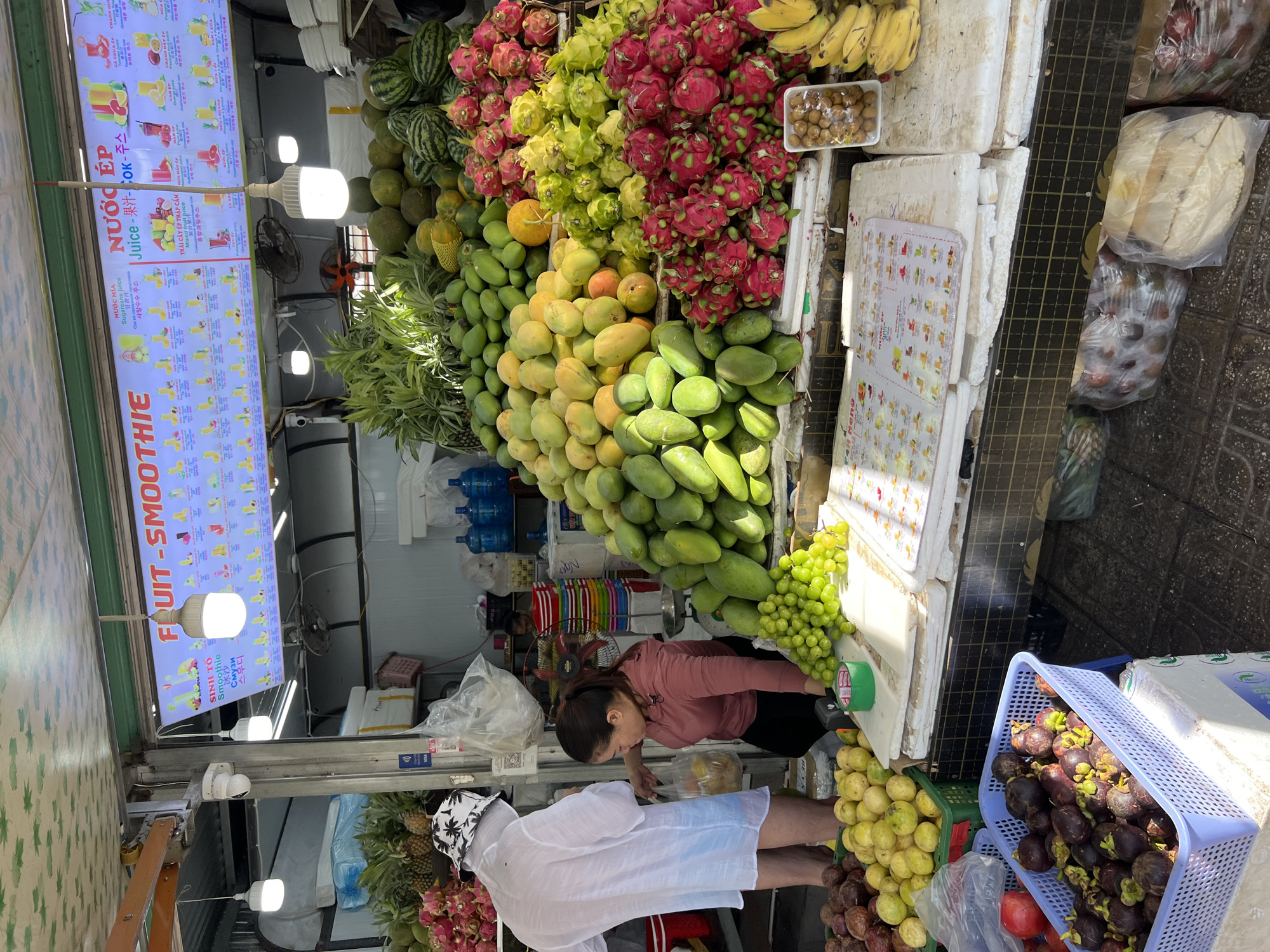The image depicts a fruit stand in Nha Trang, Vietnam, located outdoors with a partial overhead canopy. A woman in a pink long-sleeved shirt and black pants is interacting with a man wearing a white shirt and a patterned hat, who is facing away from the camera. The woman appears to be handing the man a plastic bag containing fruit.
The fruit stand is laden with a variety of fresh fruits, including green mangoes, yellow mangoes, dragon fruit, watermelons, bananas, grapes, and passion fruit. Baskets and containers are used to display the produce. Above the fruit stand, a large illuminated sign displays a menu of fruit juices and smoothies in multiple languages, including Vietnamese, English, and Korean.
In the background, there are shelves with bottled water, stacked crates, and a ceiling fan. The ground appears to be paved. The overall scene suggests a vibrant market environment selling fresh fruit and beverages.