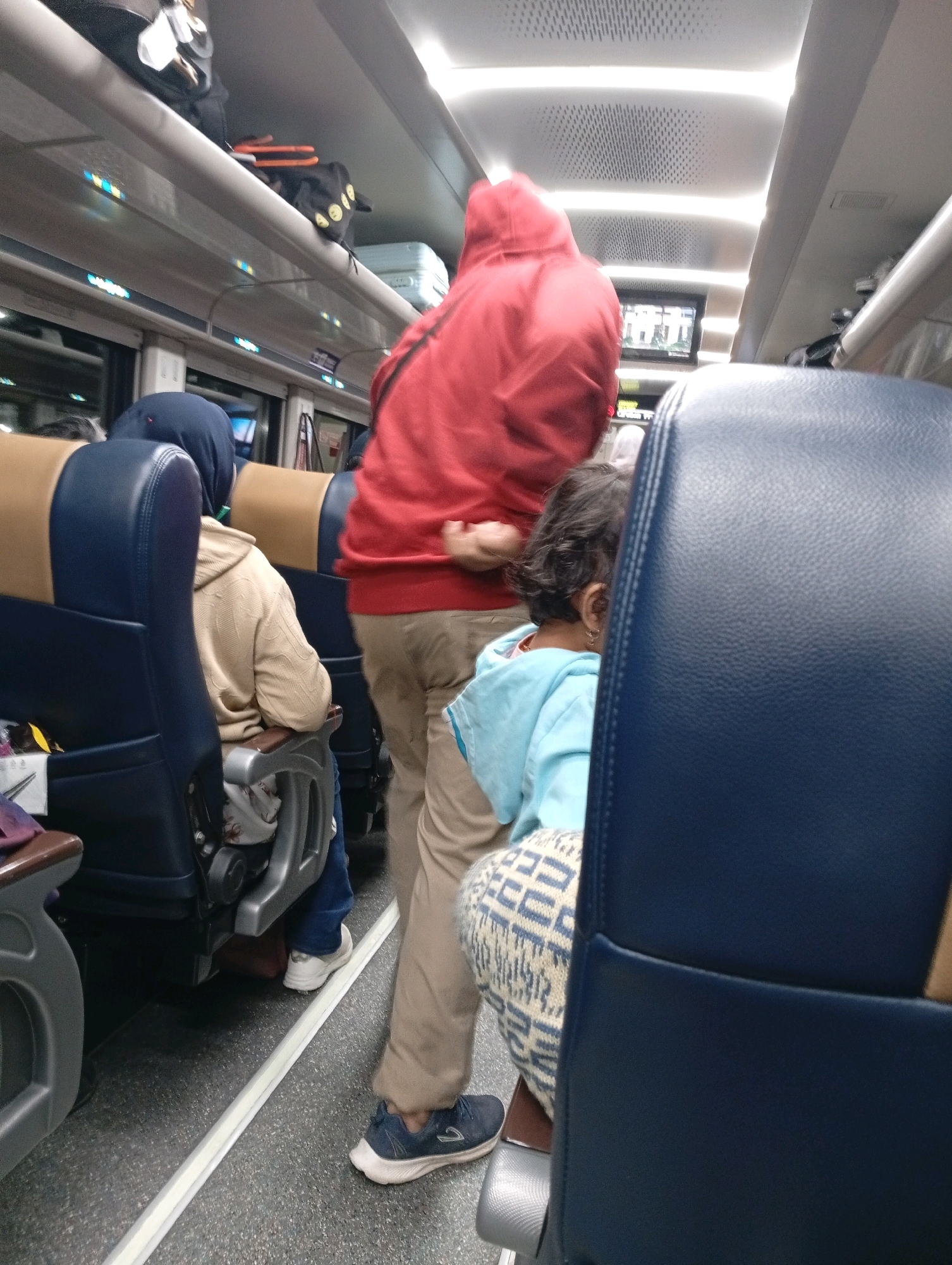 The image shows the interior of a train carriage in Cirebon, Indonesia. The scene captures passengers seated and standing. A person in a red hoodie is standing in the aisle. In front of them, a child in a light blue jacket is seated, partially obscured by a dark blue seat. To the left, a person wearing a headscarf and a beige hoodie is seated in a blue and tan seat. Overhead luggage racks contain bags. The environment appears to be a mode of public transportation.