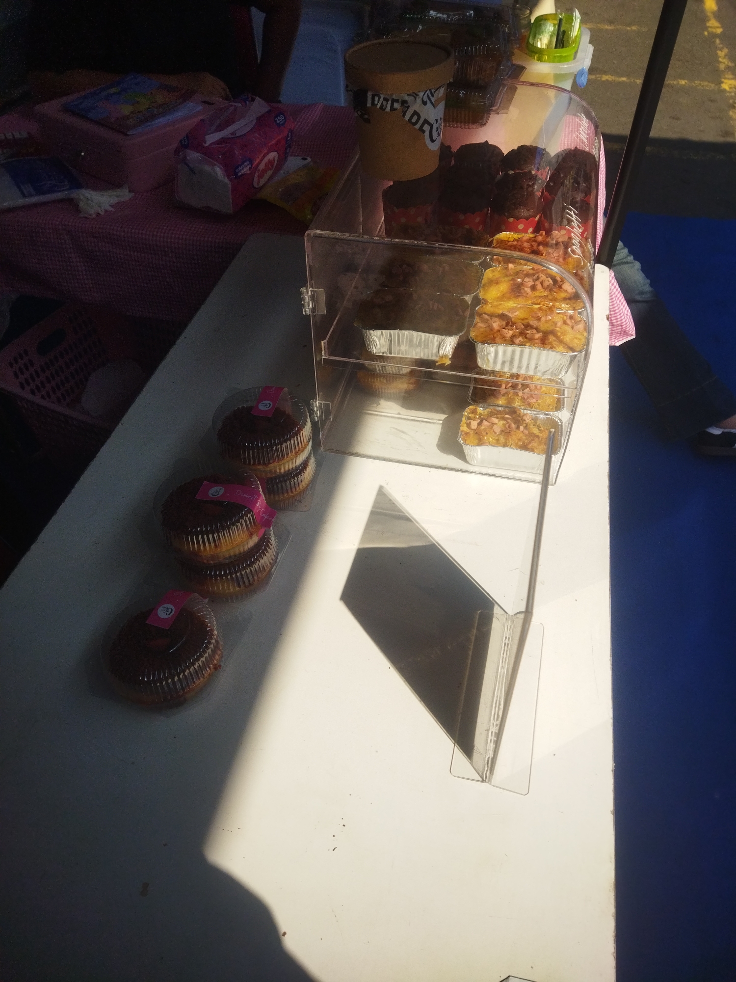 The image depicts a market stall in Giripurwo, Indonesia. In the foreground, several chocolate cupcakes are displayed in clear plastic containers with pink labels. Behind the cupcakes, a clear plastic display case contains baked goods, including what appear to be muffins or small cakes, and some foil-wrapped rectangular items with toppings. To the left, a pink and white checkered tablecloth is partially visible, on which sits a pink box, a pack of tissues, and other items. A brown paper cup with writing is also visible. The stall is outdoors, indicated by the sunlight and shadows, and a blue surface is visible in the background, possibly a table or the ground.