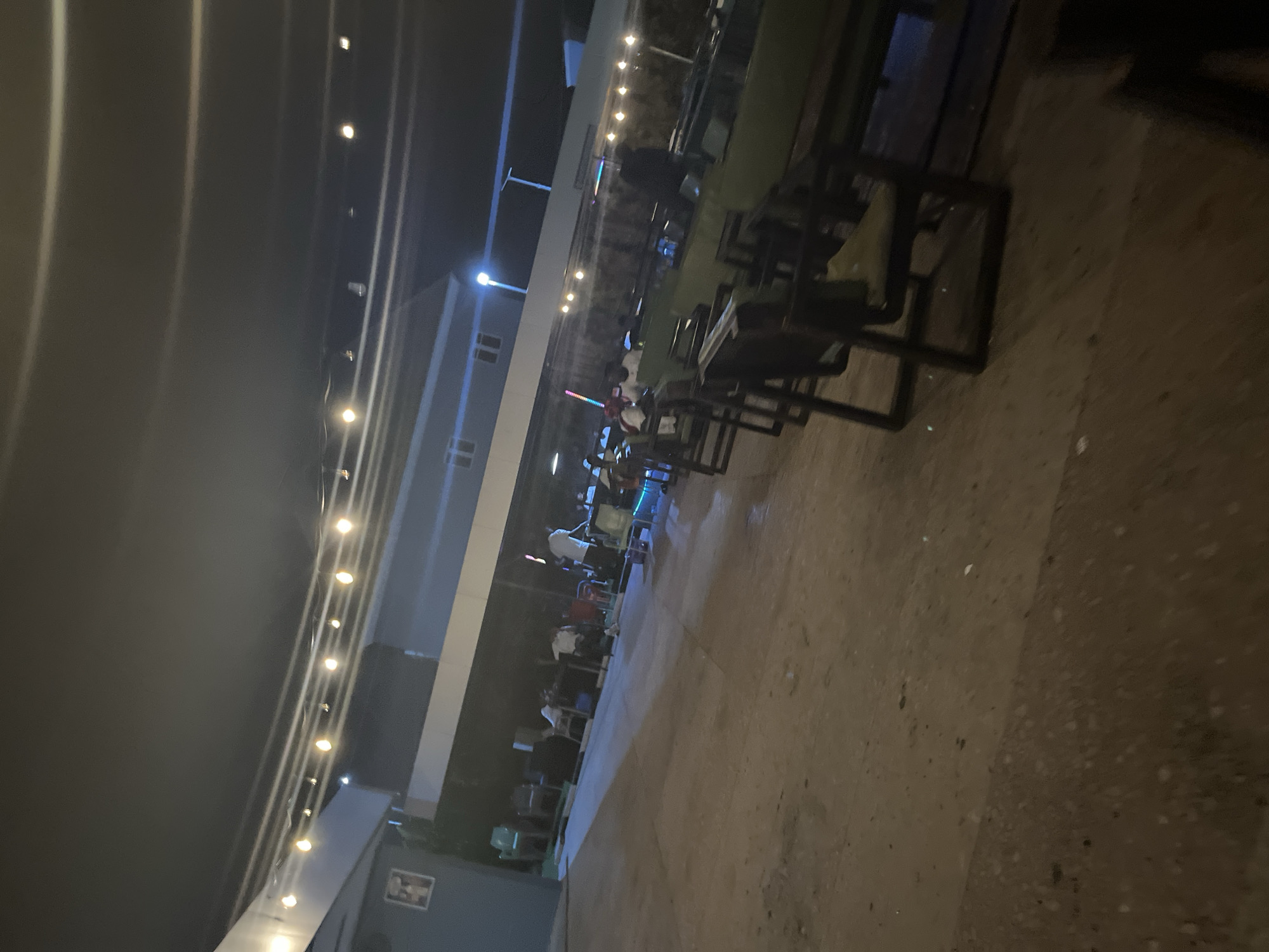 An exterior shot at night captures an outdoor seating area with string lights illuminating the scene. Several tables and chairs are visible, some occupied by patrons. A person in a white shirt stands near a table in the background. The ground appears to be paved, and a building is visible in the distance. The overall atmosphere suggests a casual gathering or dining establishment.