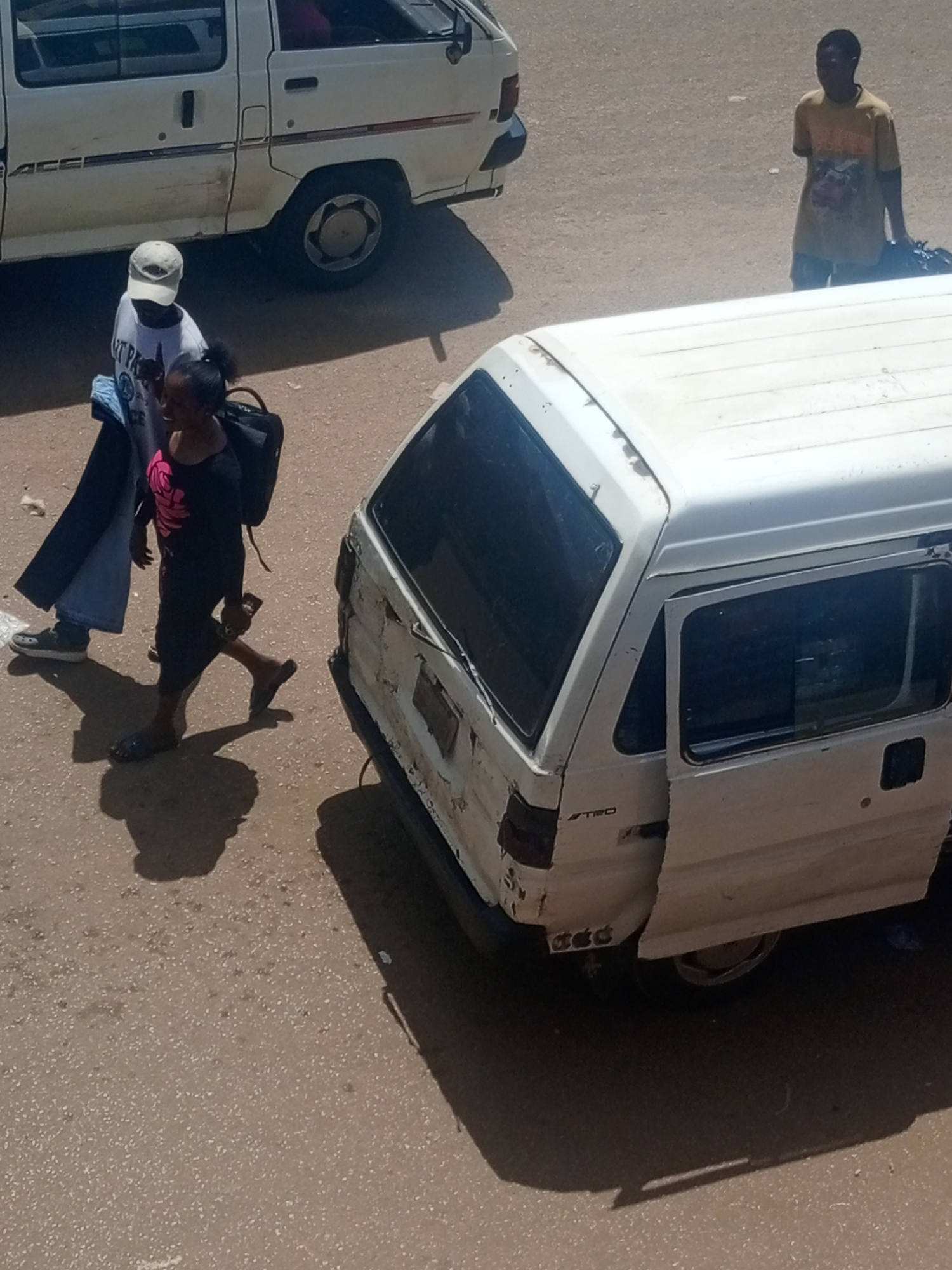 In the city of Bukuru, Nigeria, two vehicles are parked on a paved surface. To the left, a white van is parked, with its rear visible. In the center, a smaller white van is parked at an angle, with its side door open. A person wearing a yellow t-shirt and dark shorts stands to the right of the second van, holding a dark bag. To the left of the second van, two individuals are walking, one wearing a white cap, white t-shirt, and blue jeans, and the other wearing a black t-shirt and dark pants. Both are walking away from the camera and appear to be interacting. The ground is covered in a light brown, textured material, possibly asphalt or a similar road surface. The lighting suggests it is daytime, with harsh shadows cast by the vehicles and people.