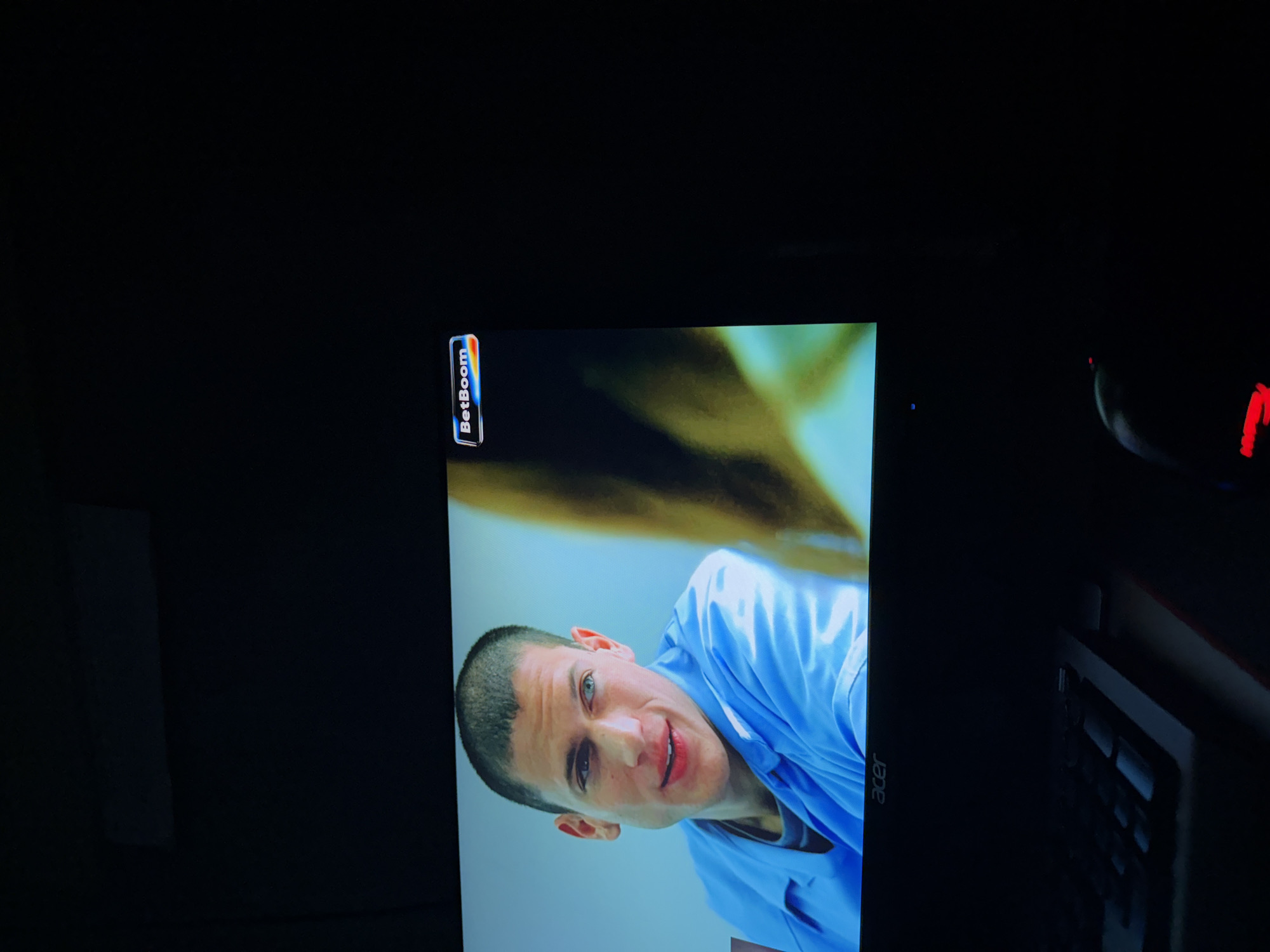The image shows a monitor displaying a man in a blue shirt. The monitor is surrounded by dark elements, possibly computer peripherals. There is a "BetBoom" logo visible in the upper left corner of the monitor. The man on screen is the subject of the display. The location is unknown.