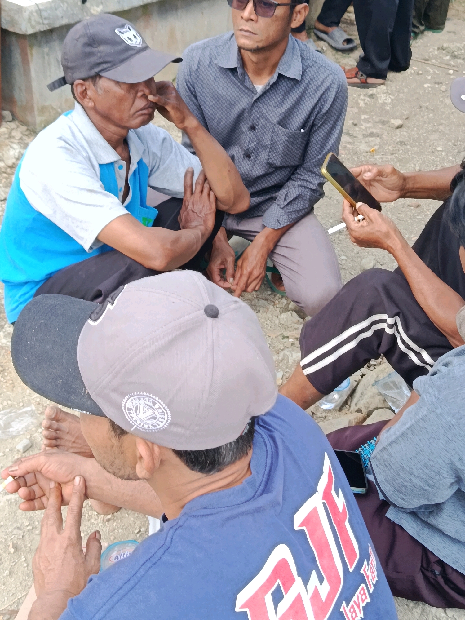 The image depicts a group of men sitting on the ground in what appears to be an outdoor setting. In the foreground, a man wearing a blue shirt with red and white lettering, and a grey baseball cap, is holding his left hand up near his face, with his index and middle fingers extended. To his left, another man in a blue and white collared shirt and a dark baseball cap is seen from the side, with his hand to his face. In the background, a man wearing a patterned grey shirt and glasses is looking down at a smartphone he is holding. Another individual, wearing black pants with white stripes, is partially visible. The ground is covered with gravel and dirt, and a plastic water bottle is on the ground. The overall impression is a casual gathering of men outdoors.
