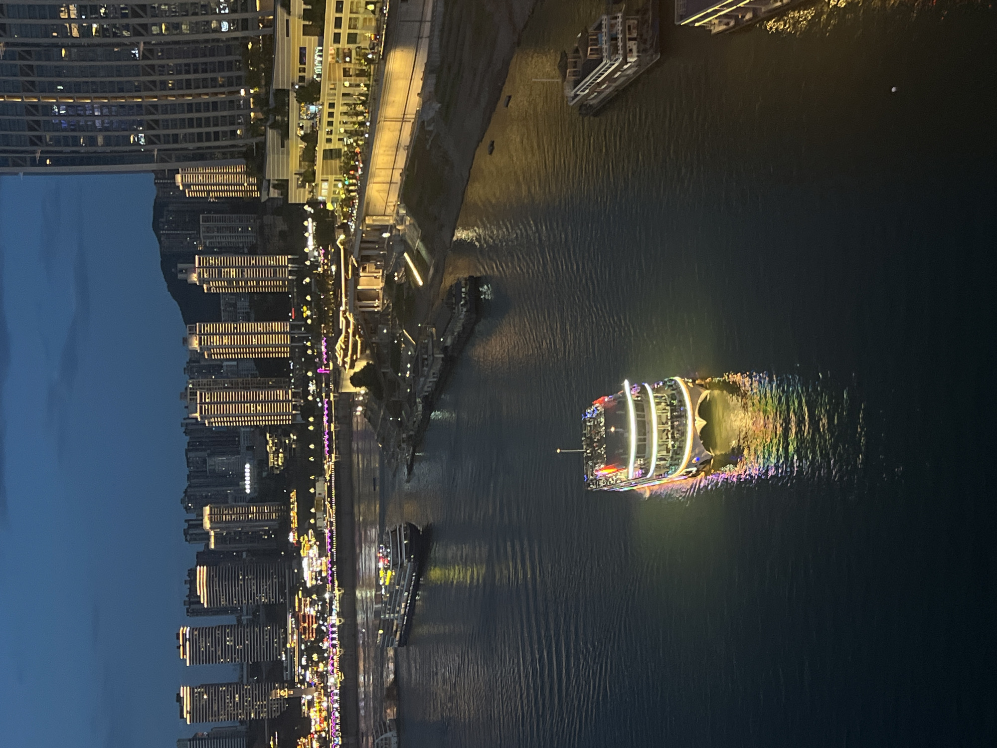 The image captures a nighttime scene of a riverfront in the Yuzhong District of China. Several boats are visible on the water, including a brightly lit passenger vessel in the foreground with colorful lights reflecting on the dark water. In the background, a cityscape with numerous illuminated buildings and a mountain silhouette can be seen. The buildings are a mix of modern high-rises and older structures, all adorned with lights, creating a vibrant urban glow against the twilight sky. The river itself appears calm, with subtle ripples on its surface. The overall atmosphere is one of a bustling, illuminated city at night, with the river serving as a central artery.