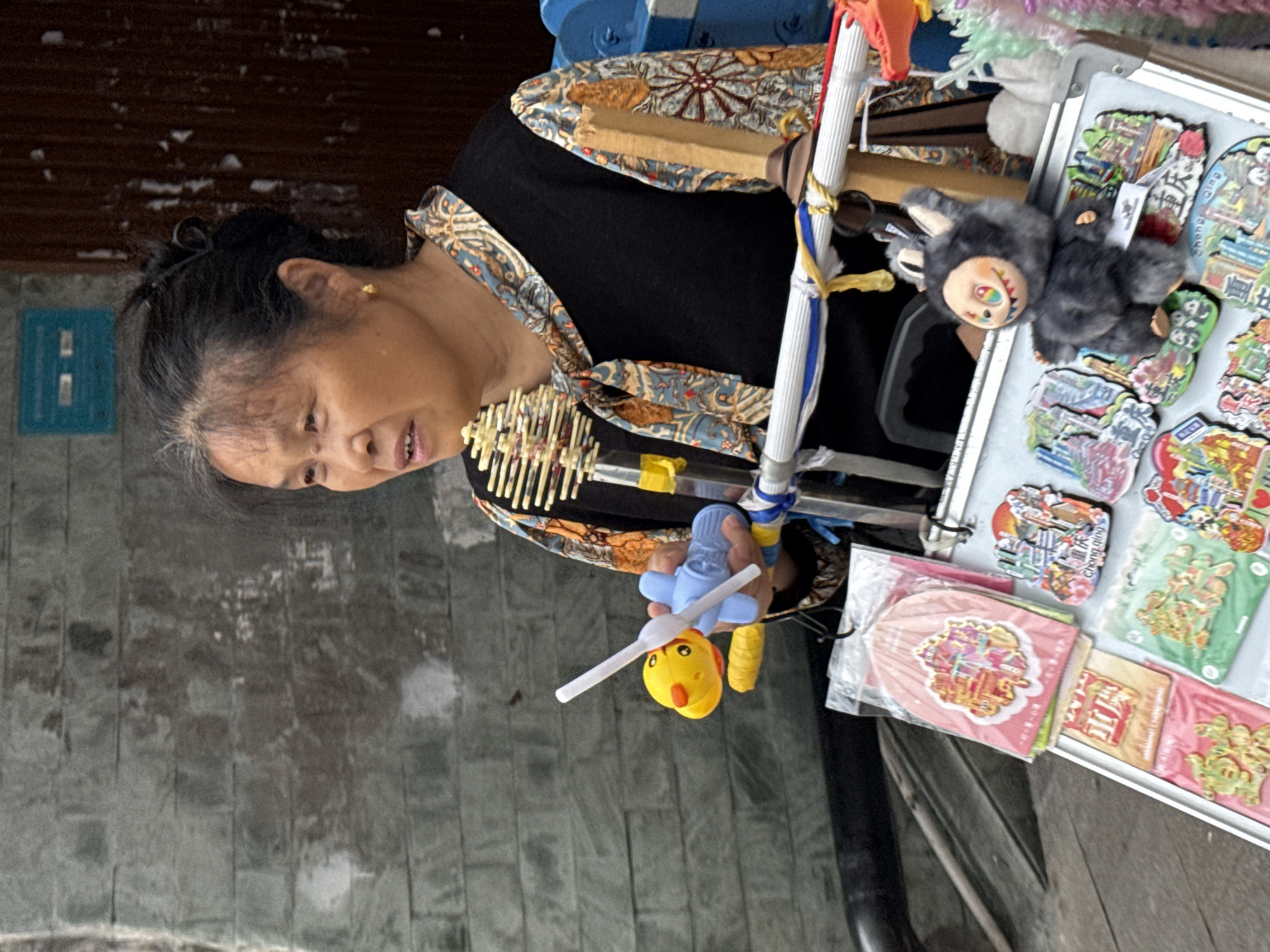 A woman is standing next to a cart filled with goods, likely for sale. The cart displays items such as stickers, a small plush toy, and other colorful trinkets. The woman is holding a toy airplane. The scene appears to be set outdoors, possibly on a street in Yuzhong District, China, given the cultural context of the goods being sold.