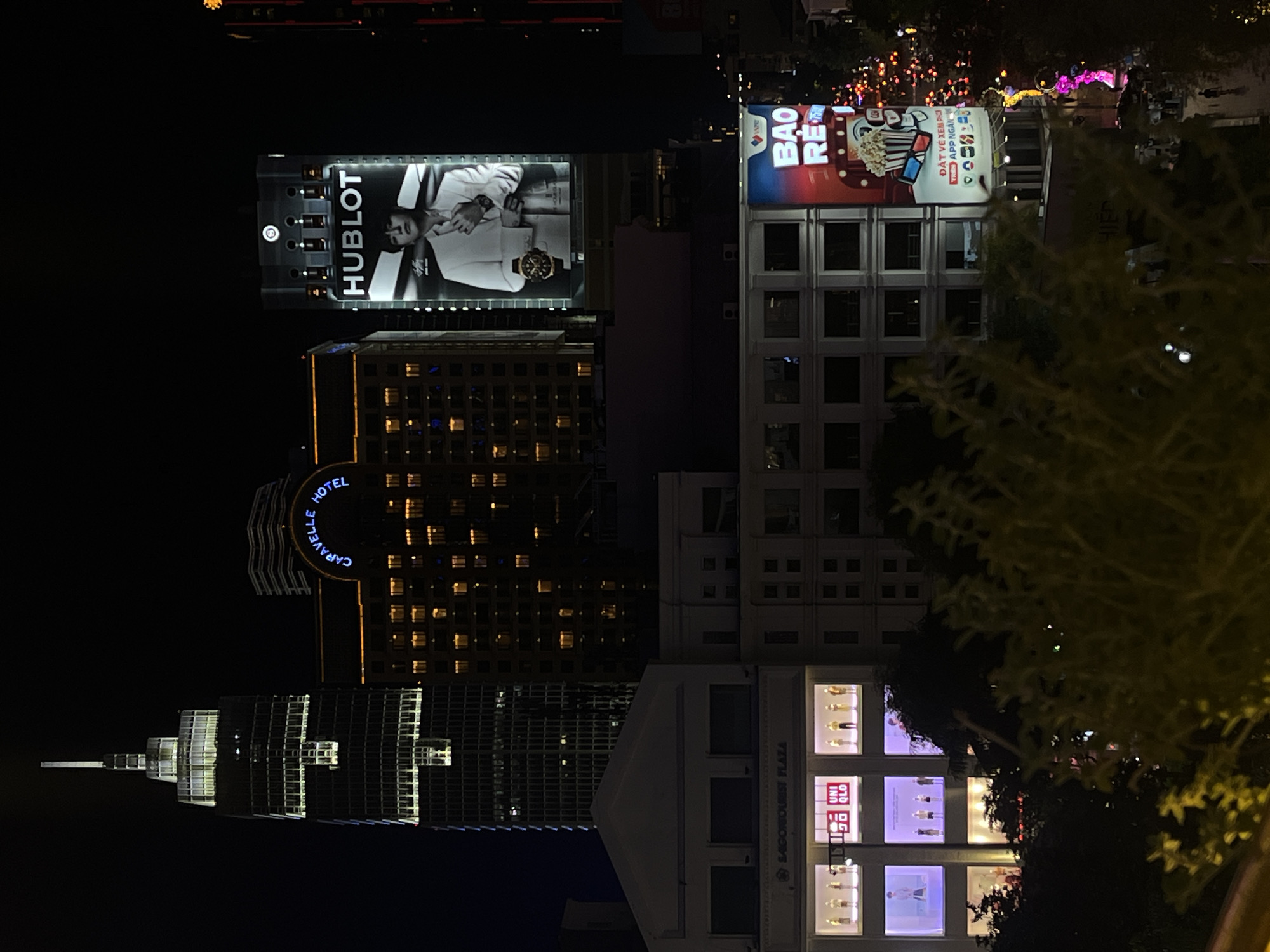 In the city of Thủ Đức, Vietnam, a nighttime cityscape is illuminated by the lights of buildings and advertisements. A tall skyscraper with a spire and numerous lit windows dominates the left side of the frame. To its right, the Caravelle Hotel is visible, its name displayed in illuminated lettering above a facade of warmly lit windows. Further to the right, a large billboard features the "HUBLOT" logo and a black and white image of a man in a white suit. Below these structures, a building with a white facade features several illuminated display windows, showcasing mannequins dressed in clothing, and a prominent "UNIQLO" logo. Another billboard, partially obscured by trees, displays colorful graphics, including a bucket of popcorn and 3D glasses, with text in Vietnamese. The lower portion of the image shows more buildings, some with lit windows, and a street with a few figures walking. Lush green foliage in the foreground partially obscures the view of the city.