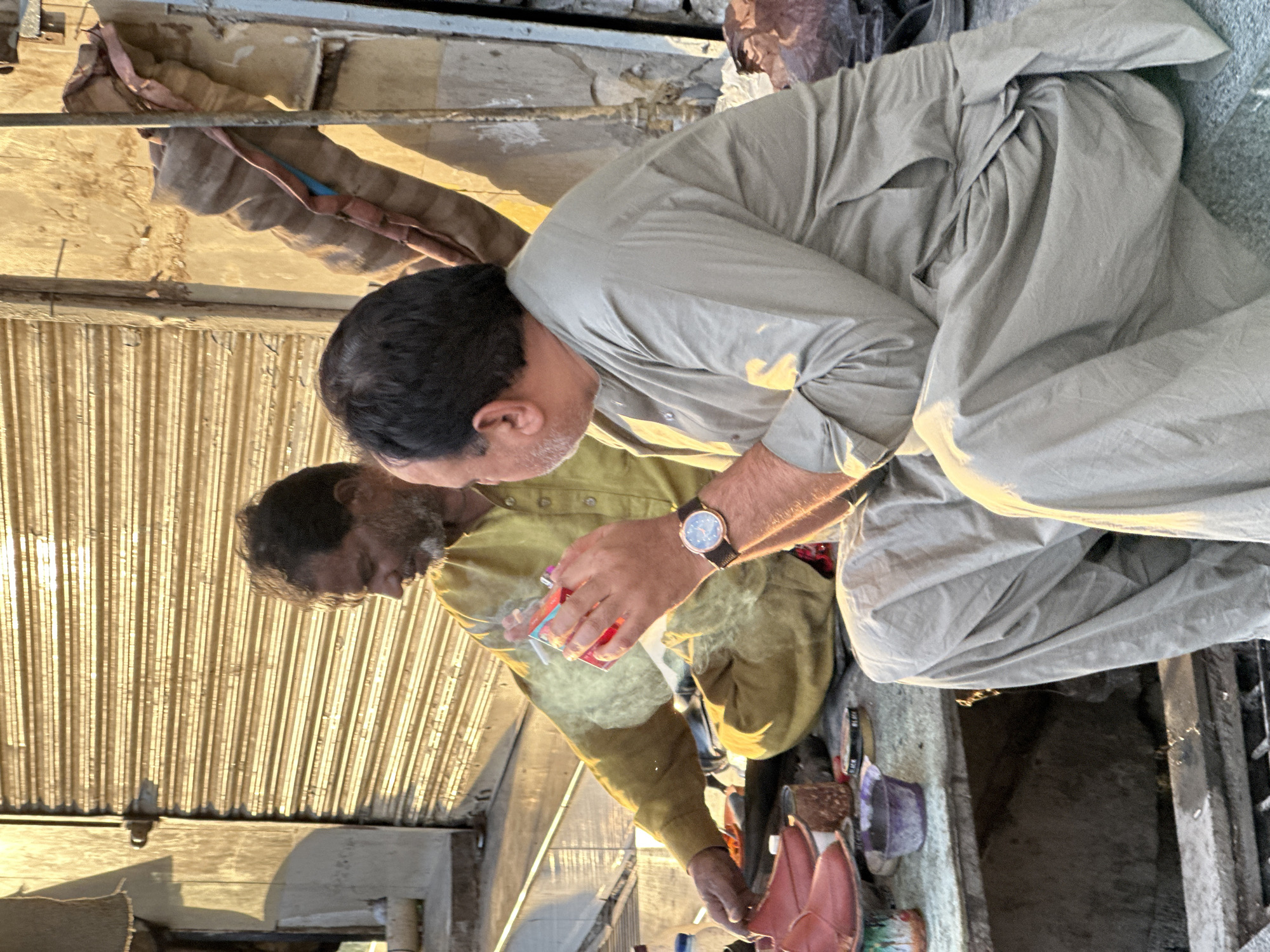 Two men are kneeling on a street in Quetta City Tehsil, Pakistan. One man is wearing a light gray traditional salwar kameez, while the other is wearing a yellow shirt. The man in the yellow shirt is holding a red item and appears to be lighting something that is emitting smoke. They are both near various containers and objects on the ground. The background includes a corrugated metal wall and various structures, indicating an urban environment.