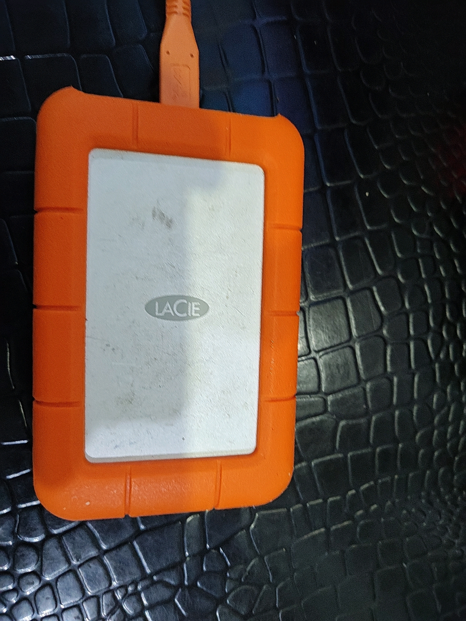 A close-up, overhead shot shows a rugged, orange external hard drive with a white surface and the "LACIE" logo on a black, textured surface. An orange USB cable is connected to the top of the hard drive. The background is a dark, patterned surface resembling alligator skin. The image appears to be taken indoors, with no discernible elements of the city of Hutatoruan 11, Indonesia.