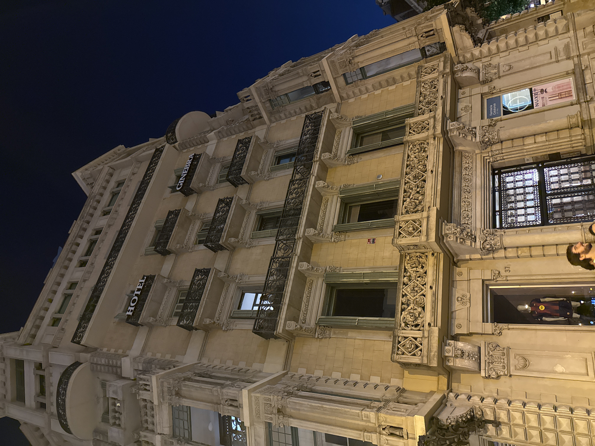 The image shows a detailed view of a historic building exterior at night in Barcelona, Spain. The building is tall, with ornate architectural features including balconies with intricate ironwork and decorative stonework. Two prominent signs are visible: one reads "HOTEL" and the other reads "GINEBRA". Several windows with green shutters are present on the facade. On the ground floor, there is an entrance with a glass door and an advertisement poster for "NUXE" and "Farmacia Catalunya Est. 1898". A mannequin wearing a soccer jersey is visible in a shop window. Two individuals are partially visible in the lower right portion of the image, with one looking towards the camera. The sky above is dark blue, indicating it is nighttime.