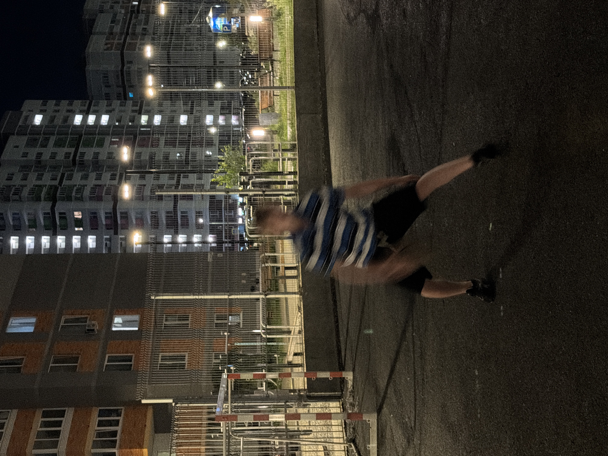 The image depicts a nighttime scene in an urban residential area, likely in Izhevsk, Russia, given the architectural style of the buildings. In the foreground, a person is in motion, captured with a motion blur, suggesting an action such as playing or exercising. The individual is wearing a horizontally striped t-shirt with blue and white bands and dark shorts, along with dark athletic shoes. The setting appears to be an outdoor court or playground, with a chain-link fence in the mid-ground. Beyond the fence, there are benches and a tree. In the background, several multi-story apartment buildings are visible, with many windows illuminated, indicating that people are inside. Streetlights are also present, casting illumination on the scene. The ground surface is dark asphalt. The overall atmosphere is that of a typical residential complex at night.