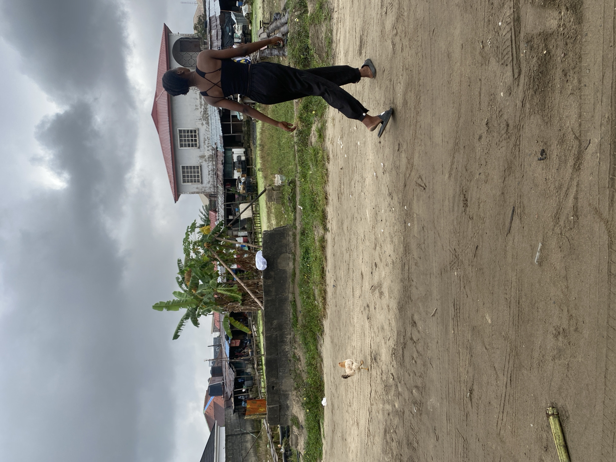 An exterior, medium shot captures a young Black woman walking away from the camera on a dirt road under a cloudy sky. She is wearing a black crop top and black pants, with black slides on her feet. To her left, a rooster walks away from the camera on the same dirt road. In the background, there is a concrete barrier with green grass and scattered white plastic bags. Beyond the barrier are various buildings, including a two-story house with a red roof, banana trees, and corrugated metal structures. The overall environment appears to be a somewhat unkempt urban or semi-urban area.