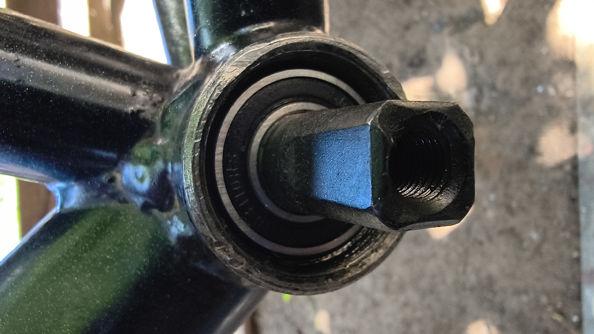 The image shows a close-up, eye-level view of a bicycle's bottom bracket assembly, specifically focusing on the spindle and bearing. The black metal frame of the bicycle is visible, with welds connecting the various tubes, suggesting it is a used or older model. The bottom bracket shell is also black and appears to be threaded, with a bearing visible inside. The spindle, which is hexagonal and black, protrudes from the center of the bearing, with visible threads at its end, indicating it is designed to accept a crank arm. The surface texture of the frame and spindle suggests it is painted metal. The background is out of focus, showing a ground surface that appears to be concrete or asphalt, with some dappled sunlight and shadows, possibly indicating an outdoor setting. There are no people or animals present in the image. The scene is forensic in nature, documenting a mechanical component of a bicycle. The location in Mikhaylovka, Russia is not discernible from the image itself.