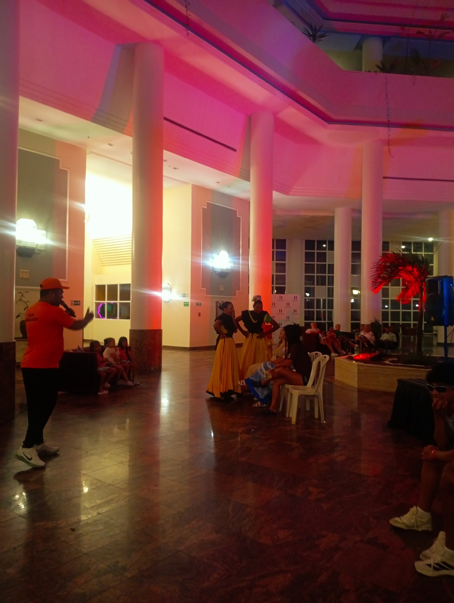 The image shows an indoor performance space, possibly a hotel lobby or convention center, in Pedro González, Venezuela. The location is identifiable by the presence of "H" logos on windows. In the foreground, a man in an orange shirt and cap stands on a polished floor, speaking into a microphone. Three women in traditional yellow and black dresses are positioned centrally, seemingly prepared for a dance performance. A person sits on a white plastic chair near them. A small audience is seated in the background. Tall, white pillars support the ceiling, which is illuminated with a pinkish hue. A potted palm-like plant is also present on the right side of the background.