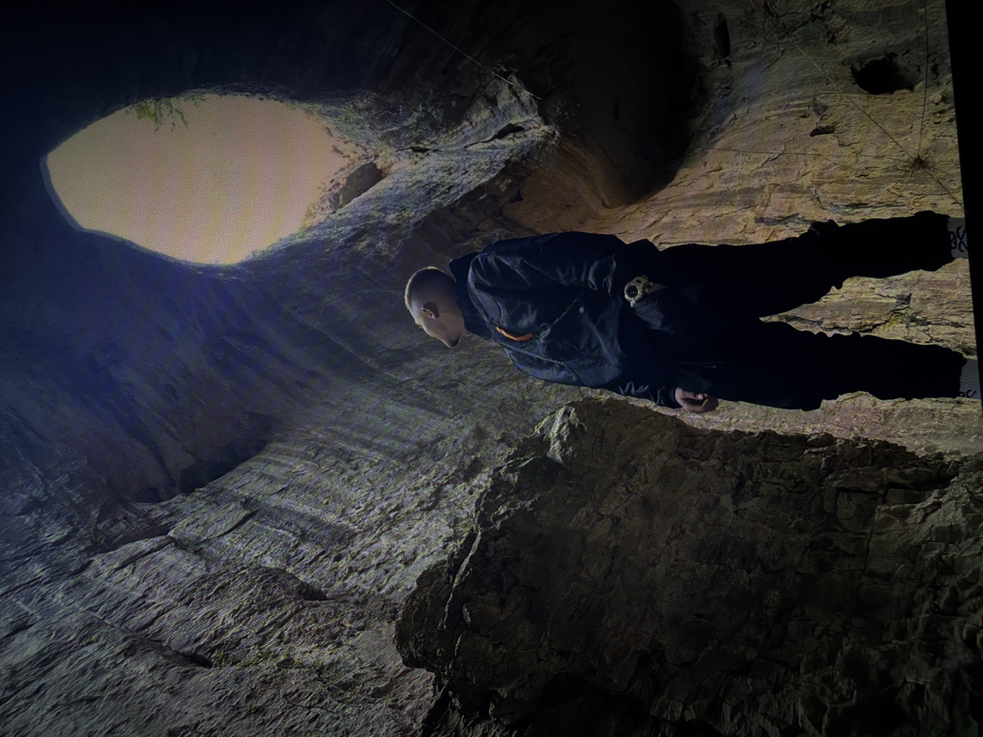 A low-angle shot shows a person standing in a cavern, looking up. The person is wearing a dark jacket with orange accents and dark pants, and is looking towards the top of the cavern, where a large opening allows sunlight to enter. The cavern walls are rough and textured, appearing to be made of rock. The light from the opening creates a bright, hazy effect.