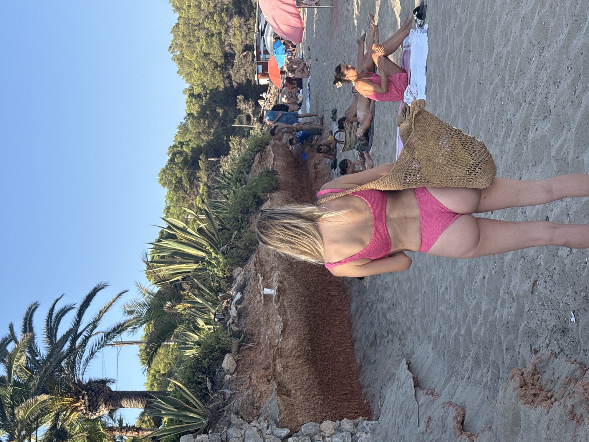 The image depicts a beach scene in Santa Eulària des Riu, Spain, under a clear blue sky. In the foreground, a woman from behind is wearing a pink bikini and carrying a woven bag. The beach is sandy, with some rocky, eroded cliffs to the left, adorned with agave plants and palm trees. In the background, several people are sunbathing on towels, some under colorful umbrellas. A small beach structure, possibly a bar or kiosk, is visible further down the beach. The overall atmosphere suggests a warm, sunny day at a popular coastal destination.