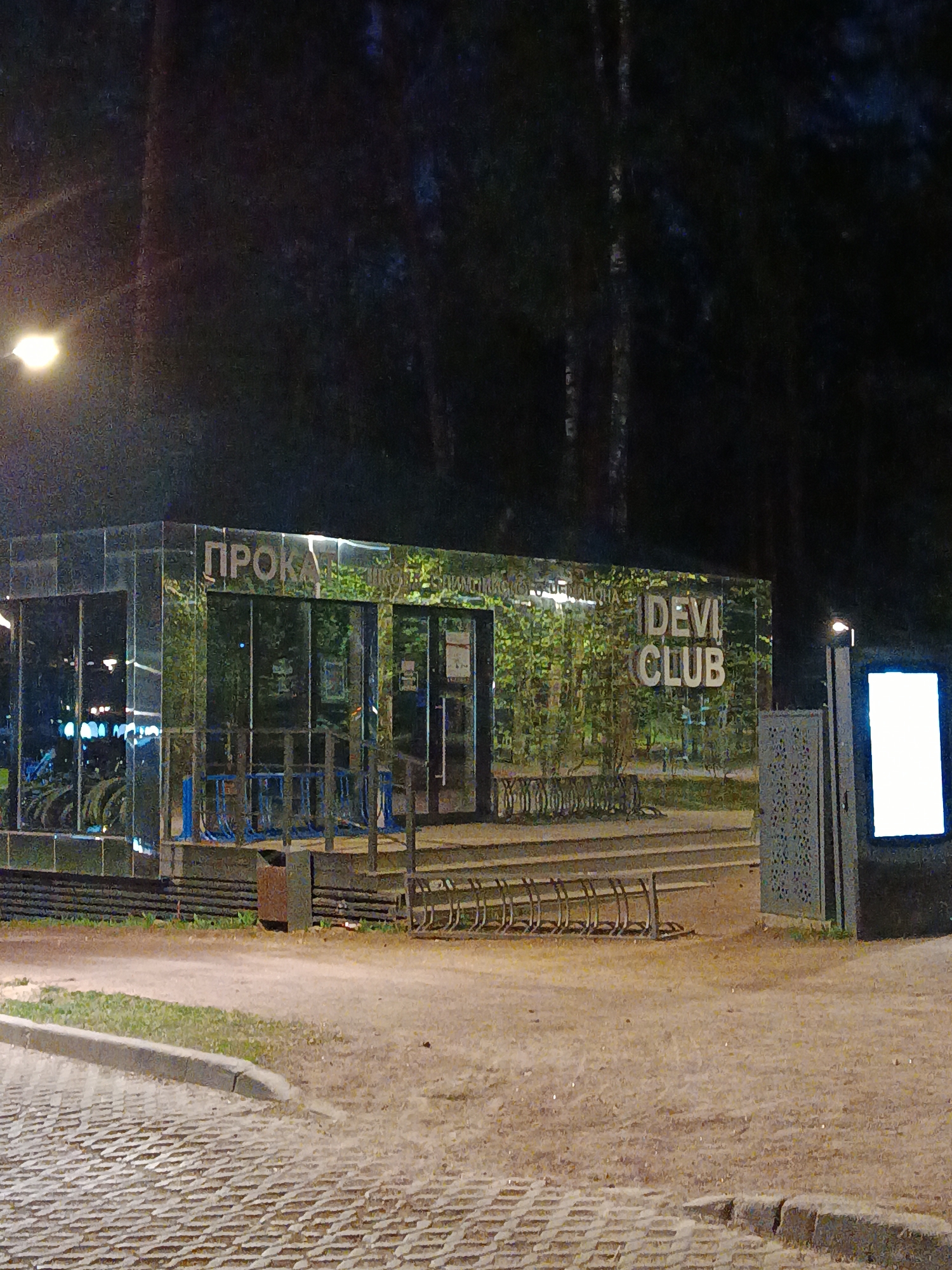 The image depicts an exterior view at night of the "Devi Club" in Barvikha, Russia. The foreground consists of a paved walkway leading to a dirt area where there are bicycle racks. To the left stands a reflective, mirrored structure with the word "ПРОКА" (Rental) visible on top. Further to the right is a sign that reads "DEVI CLUB". A backlit digital sign is on the right-hand side. A street lamp illuminates the area. The background consists of dark trees and foliage.