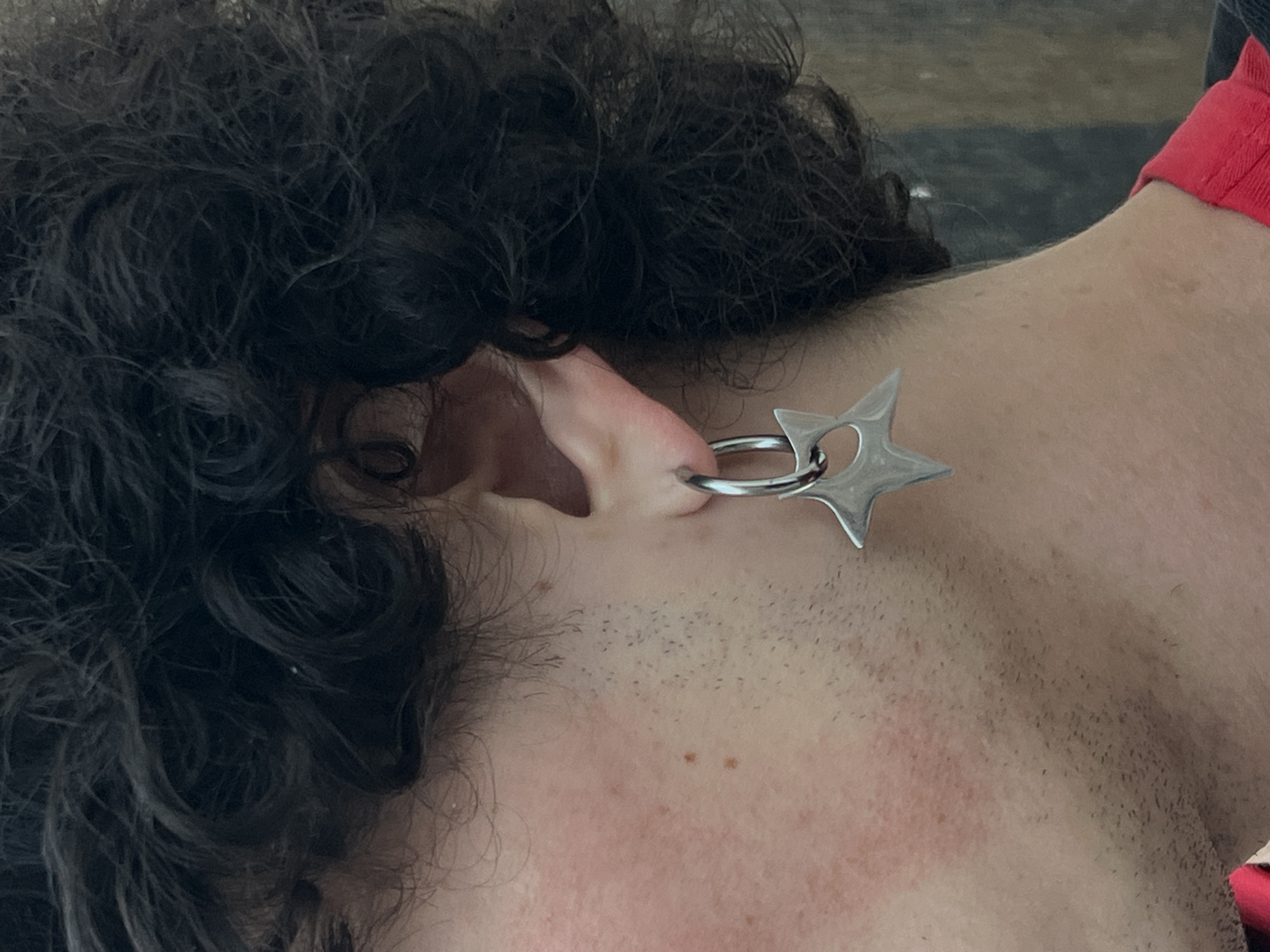 A close-up shows a person with dark, curly hair and fair skin. The person's ear is pierced with a silver ring earring that has a star-shaped charm attached. The person is wearing a red shirt with a ribbed collar. The background is blurry and indistinct. The location is not discernable from the image.