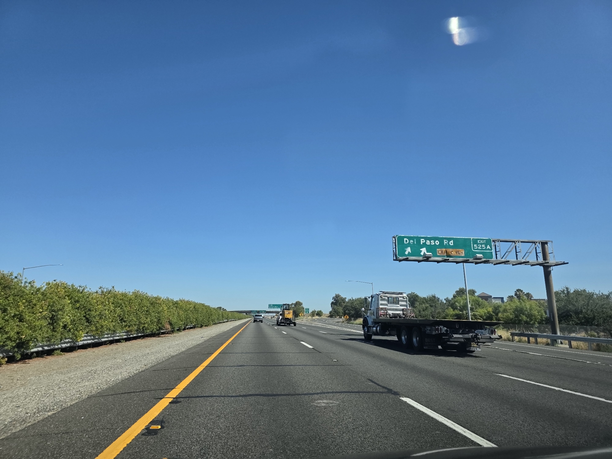 A flatbed truck drives on the right lane of a multi-lane highway under a bright, clear blue sky. The truck, with a white cab and a dark flatbed, is the most prominent object. In the distance, a yellow excavator is visible on the left side of the highway. A green overhead highway sign indicates "Del Paso Rd EXIT 525A" with an arrow pointing left. The highway is lined with trees and bushes on the left and right sides. The road surface is asphalt with clear lane markings. The scene appears to be on a sunny day, likely in the city of Sacramento, California, United States of America.