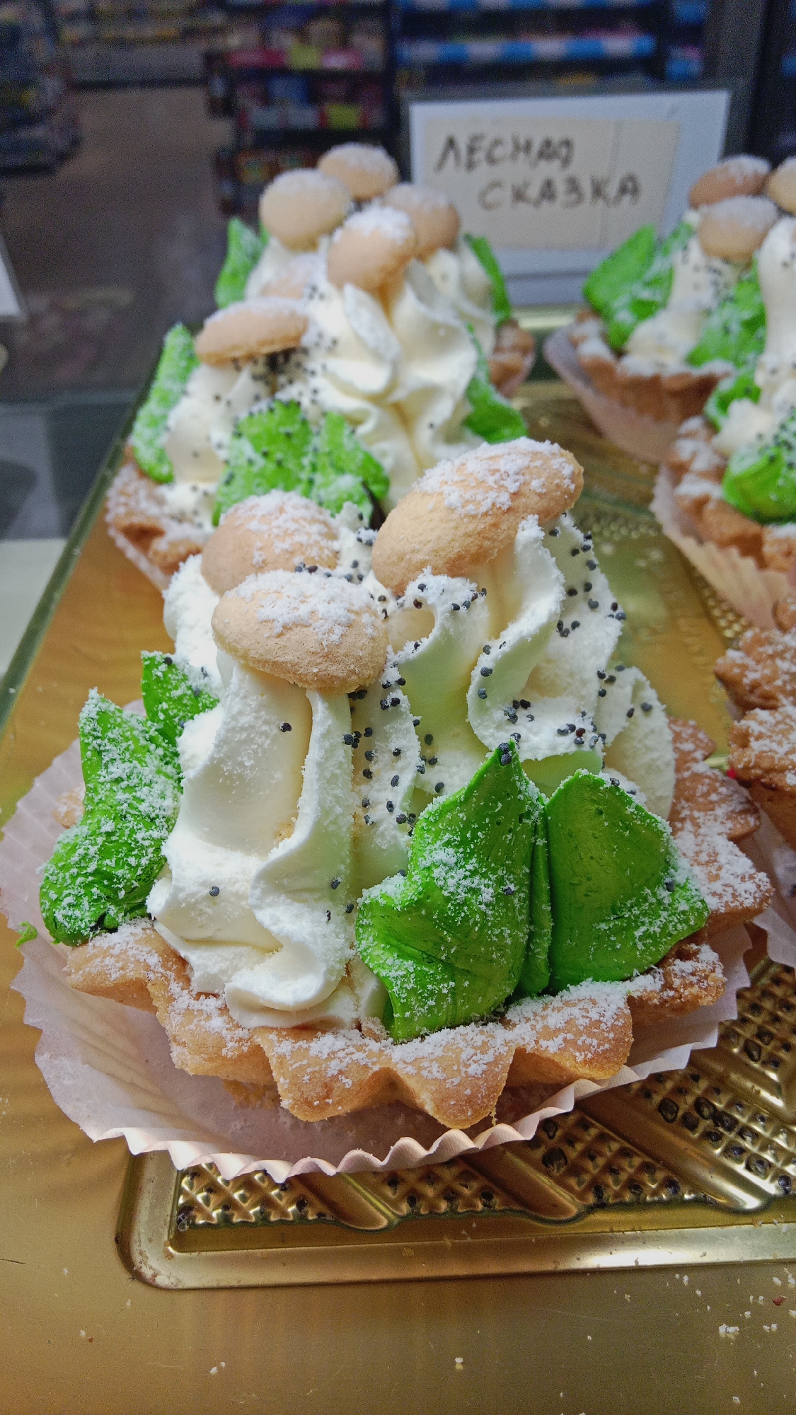 The image shows a close-up of several cream-filled tarts arranged on a golden tray. Each tart is in a fluted, paper-lined pastry shell. The tarts are elaborately decorated with swirls of white cream, green frosting shaped like leaves, small, oval-shaped cookies resembling mushrooms, and sprinkled poppy seeds. The white cream is dusted with what appears to be powdered sugar or shredded coconut. In the background, out of focus, is a sign with Cyrillic writing that reads "ЛЕСНАЯ СКАЗКА," which translates to "Forest Tale." The tarts are likely for sale in a bakery or pastry shop. The setting is indoors, possibly a retail display. Based on the sign, this bakery is located in Petropavl, Kazakhstan.