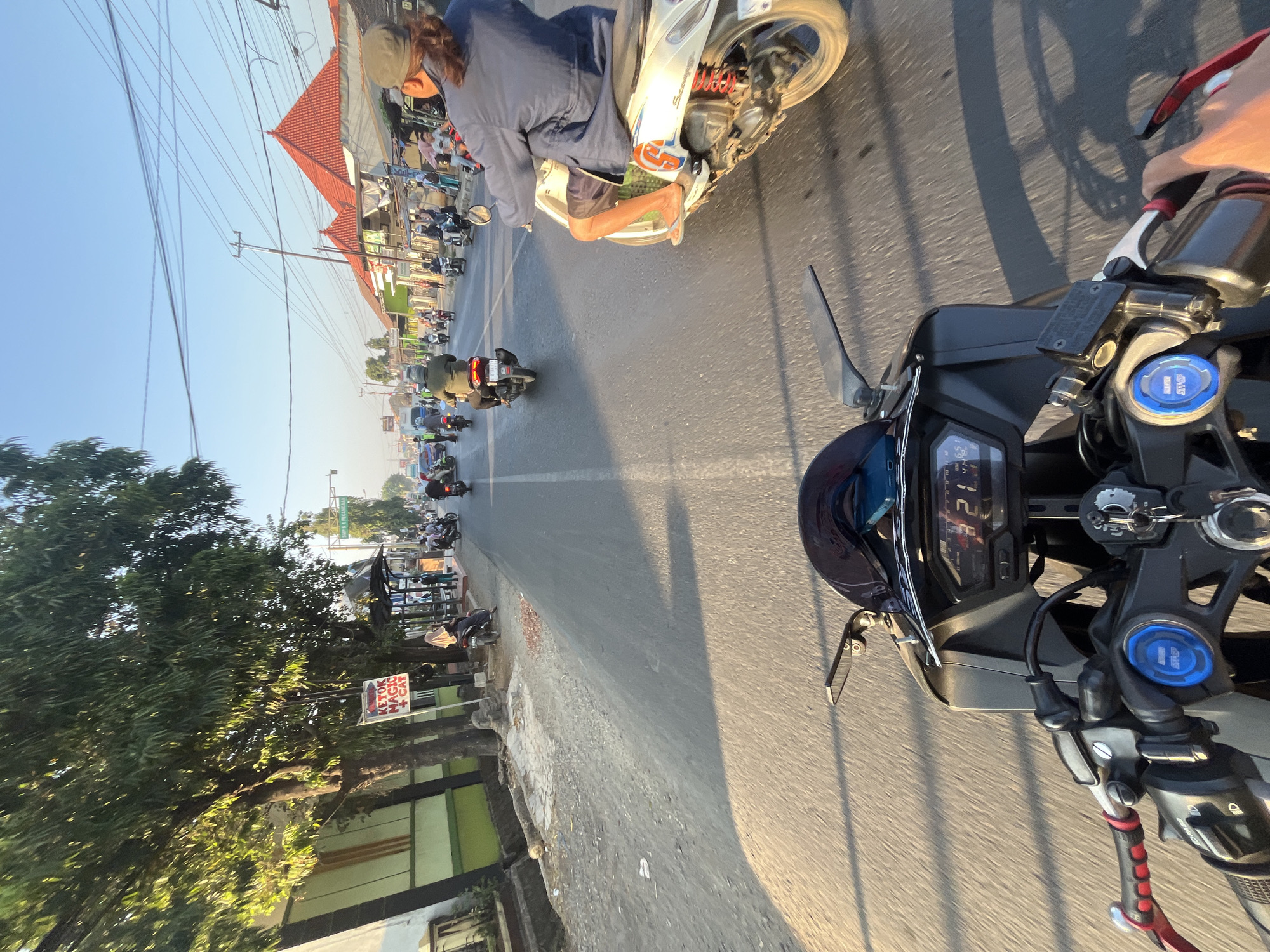 This is a first-person perspective photograph taken from a motorcycle traveling on a paved road in Kalinyamatan, Indonesia. The time of day appears to be late afternoon, indicated by the long shadows cast across the road and the warm, golden sunlight illuminating the scene.
The foreground is dominated by the handlebars and dashboard of the motorcycle from which the photo is taken. The dashboard displays a digital speedometer reading "21". The road surface is asphalt, with visible lane markings and some debris scattered along the side.
In the middle ground and background, traffic flows in both directions. Numerous motorcycles and a few cars are visible. Some motorcycles carry one or two riders, many of whom are wearing helmets. One motorcycle in the right lane has a rider wearing a dark blue jacket and shorts. A white scooter with red shock absorbers is positioned in the right lane, with its rider's legs visible.
Along the left side of the road, there are trees, a building with a green and white facade, and a sign that reads "KETOK MAGIC + CAT". There is also a bus stop shelter. Along the right side, buildings with red tiled roofs are visible, interspersed with smaller structures and stalls. Utility poles with a network of wires are present above the road. The sky is clear and blue. The overall scene depicts a typical urban streetscape in Indonesia with moderate traffic and pedestrian activity.