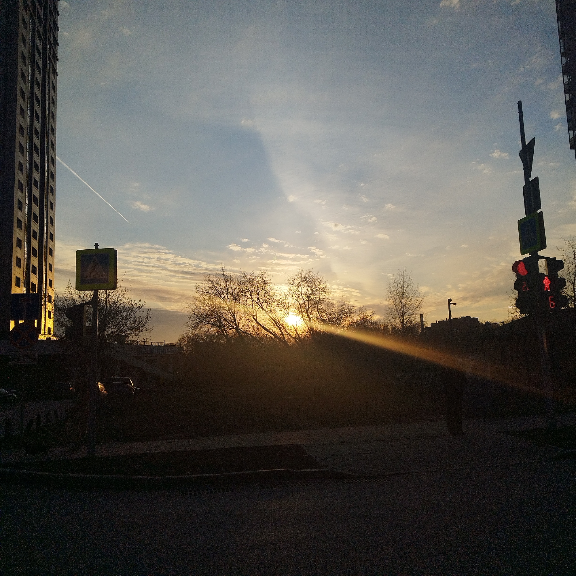 The image captures an urban street scene at either sunrise or sunset, characterized by strong backlighting from the low sun. The sun, partially obscured by bare trees and bushes in the middle ground, casts a bright, warm glow across the sky and creates a prominent lens flare or sunbeam extending diagonally from the center-right. The sky transitions from light orange/yellow near the horizon to a pale blue at the top, with scattered, thin clouds. A long, white airplane contrail streaks across the upper-left sky.
On the far left, a tall, multi-story building with numerous windows stands silhouetted, its left facade catching some of the golden light. On the far right, another building is partially visible. In the foreground, the street and sidewalk are almost entirely in shadow, appearing dark and indistinct, though faint outlines of parked cars can be seen.
Traffic infrastructure is visible: a yellow square pedestrian crossing sign with a black symbol is positioned left of center, while a traffic light for pedestrians is on the right. The pedestrian traffic light shows a red "don't walk" symbol and a digital countdown displaying the number "6" (and possibly "2" above it). The overall impression is a tranquil yet stark view of a city preparing for or winding down from the day, with the natural light creating dramatic contrasts. No specific human activity is clearly discernible, but the urban elements suggest a populated area.