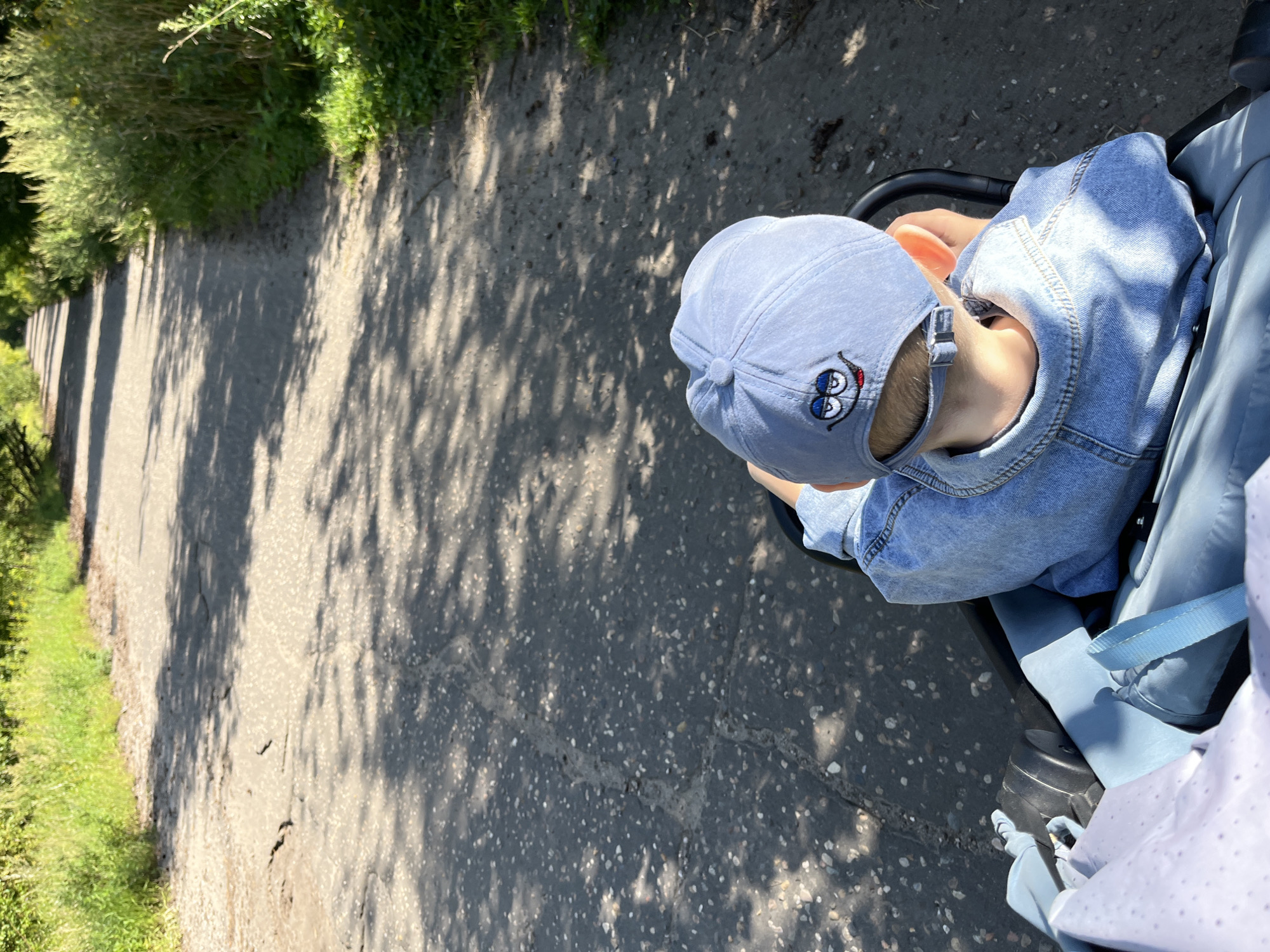 A young child, seen from behind, is seated in a stroller on a cracked asphalt path. The child wears a light blue denim jacket and a light blue baseball cap with a cartoon face embroidered on the back. The path is lined with tall green grass and trees, casting dappled shadows on the ground. The scene appears to be outdoors, possibly in a park or rural area. The location is identified as Balashikha, Russia.