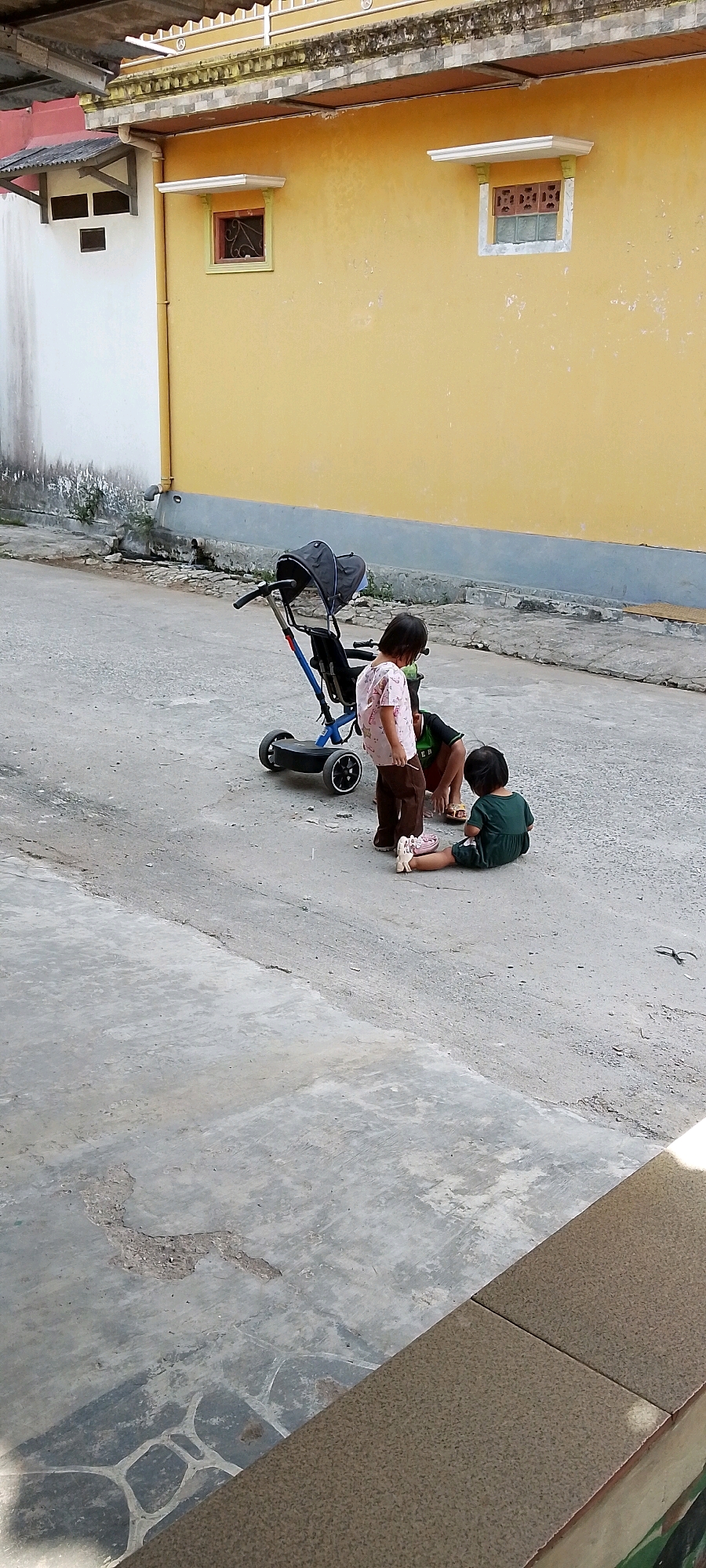 In the city of Satriamekar, Indonesia, a scene unfolds on a paved outdoor area adjacent to a building with a yellow facade. Three children are present. One child, wearing a light-colored, patterned shirt and brown pants, stands near a blue and black stroller. Another child, wearing a dark green dress, sits on the ground facing away from the viewer, with their legs extended. A third child, mostly obscured by the other two, is also on the ground. The stroller is positioned in the middle ground, and the children appear to be interacting with each other or the stroller. The environment shows a concrete ground with some cracks and debris, and the building wall behind them exhibits some weathering.