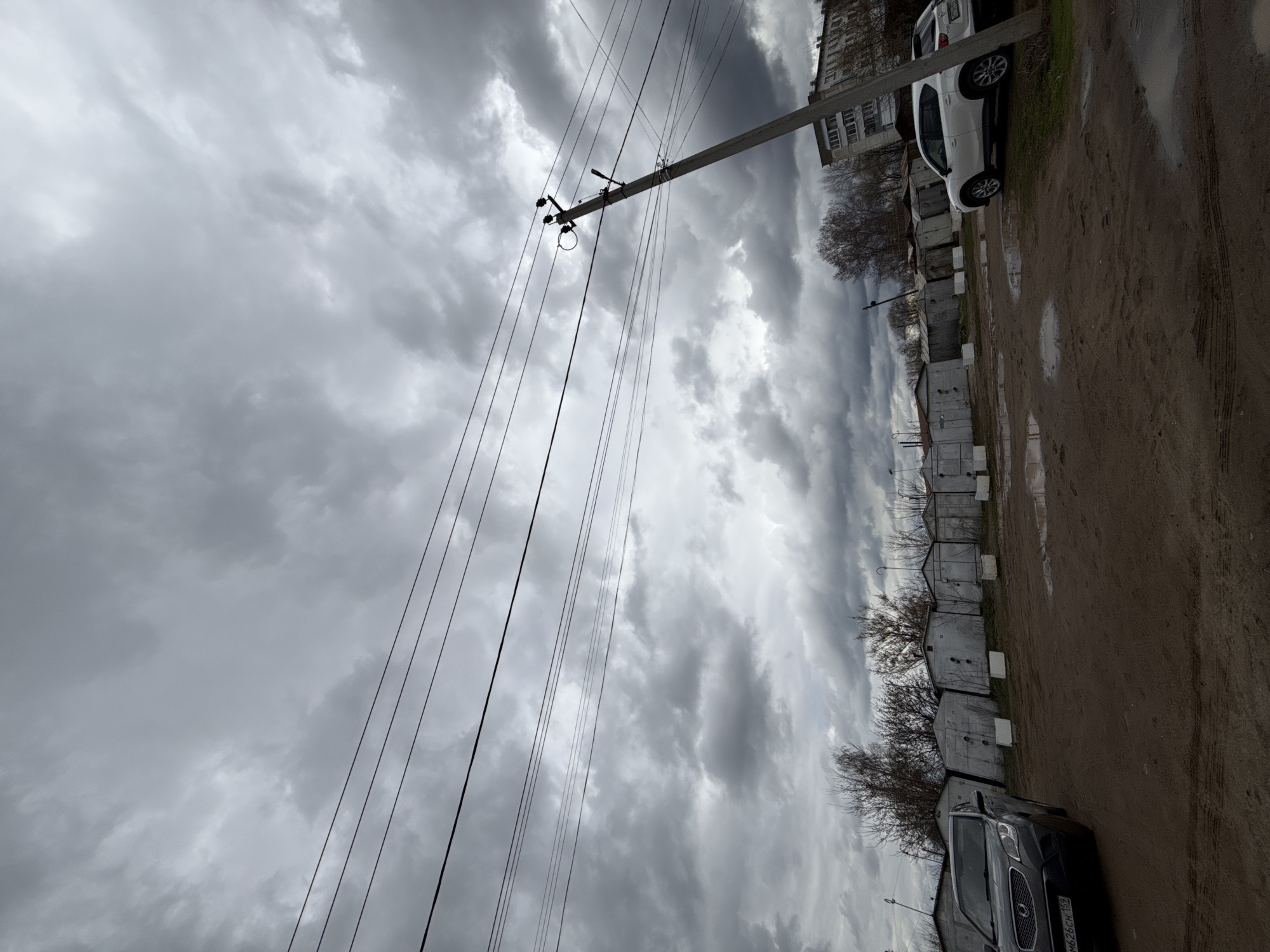 The image, taken in Chaikovsky, Russia, captures a gloomy, overcast day over a functional outdoor environment. The sky is dominated by heavy, dark grey clouds, indicating an impending storm or recent rainfall, casting a diffused and subdued light over the scene.
In the foreground and midground, an unpaved dirt road stretches across the lower portion of the frame. It appears muddy and uneven, with visible puddles and tire tracks. Along the right side of this road, a long row of simple, utilitarian garages or storage units, likely constructed from concrete blocks, extends into the distance. Beyond these structures, a multi-story residential building, an apartment block, is visible in the far background.
Two vehicles are present: a white SUV or hatchback is parked on the dirt road towards the right side of the frame, partially on a grassy verge. In the lower left foreground, a dark-colored SUV or crossover is also parked on the dirt road, displaying a visible Russian license plate: "m626CH 152".
A prominent utility pole, featuring a street lamp fixture and numerous power lines, diagonally traverses the left half of the image. Sparse, bare trees or bushes, devoid of foliage, are scattered along the right side of the road, between the garages and the distant building, suggesting late autumn, winter, or early spring. No specific activity is taking place; the image presents a static snapshot of an urban edge or industrial area under a somber sky.