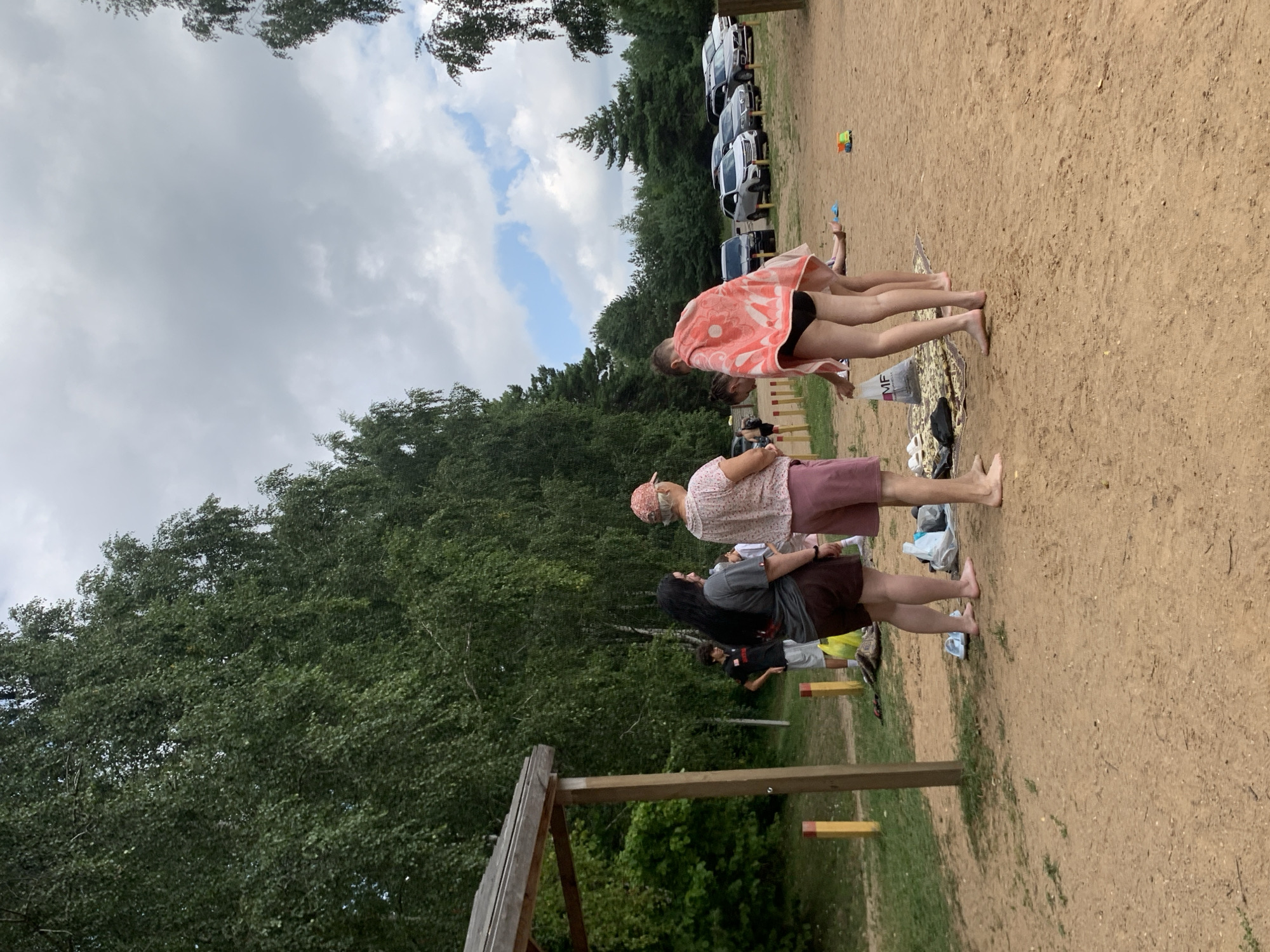 In the city of Грозаўскі сельскі Савет, Belarus, a group of people are gathered on a sandy area with trees in the background. In the foreground, four individuals are visible. Two women, one with long dark hair and wearing a grey top and brown shorts, and another in a pink cap and pink shorts, stand near each other. A third person, wearing a pink towel draped over their shoulders and black bikini bottoms, is bending down. A fourth person, partially obscured, is also in the vicinity. To the left, a wooden structure with a roof stands near a grassy area with yellow and red posts. In the middle ground, several cars are parked in a line. The overall environment appears to be a recreational area, possibly a beach or a park with a parking lot. The sky is overcast with some patches of blue.