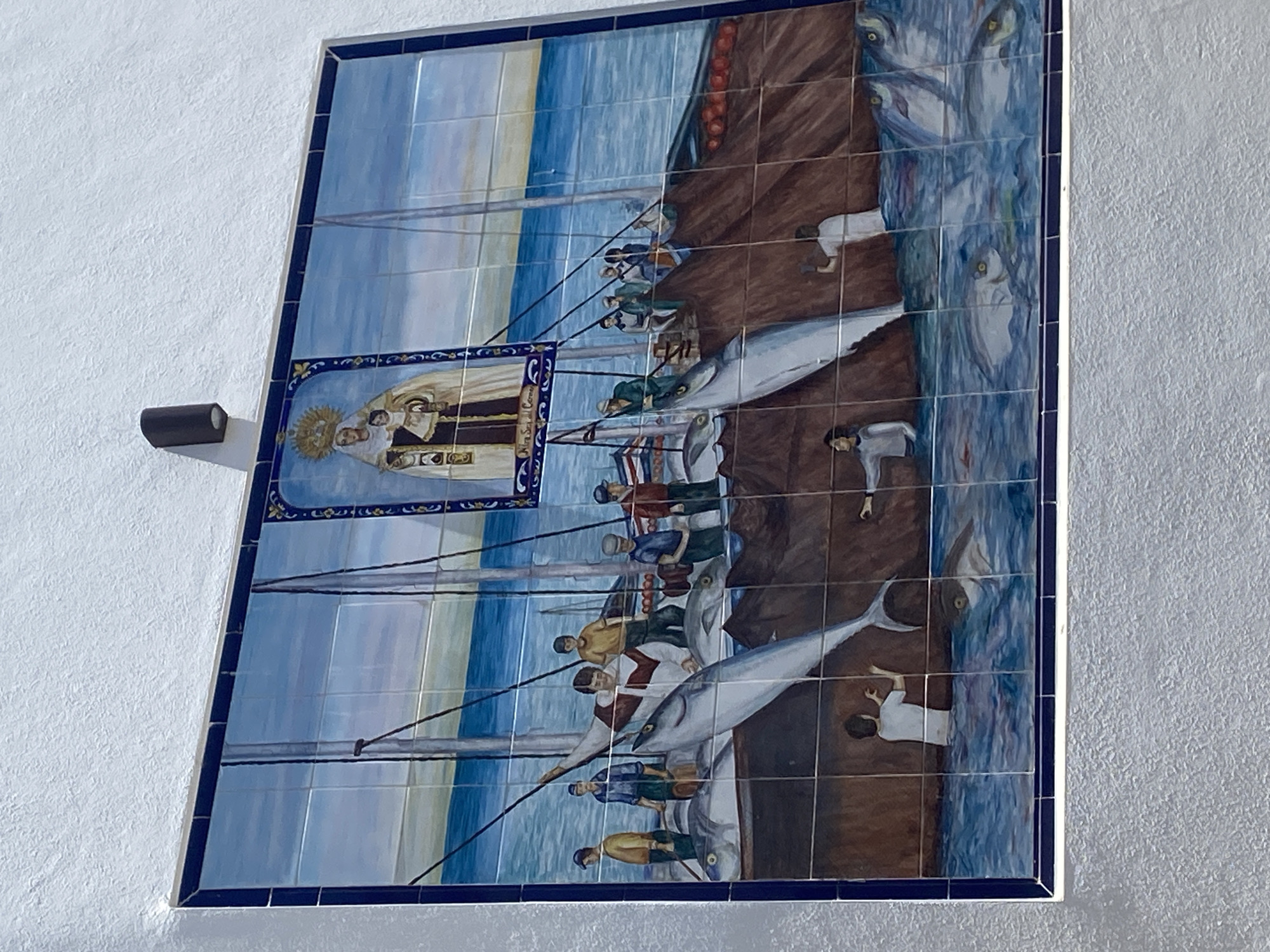 This is a detailed description of a ceramic mural located in Conil de la Frontera, Spain, depicting a scene related to fishing and religious veneration.
The mural is composed of numerous ceramic tiles, arranged in a grid pattern and framed by a dark blue border. The artwork portrays a bustling fishing scene at sea, with a central focus on several large fish, possibly tuna, being brought aboard a boat. Several men, dressed in simple, historical-looking clothing, are actively engaged in the fishing operation. They are shown pulling ropes, handling the fish, and standing on the deck of the boat. The water is depicted with varying shades of blue, suggesting depth and movement.
Above the fishing scene, within a smaller, ornate frame, is an image of a religious figure, likely the Virgin Mary, holding a child. This religious iconography is placed centrally in the upper portion of the mural, implying a connection between the fishing activities and a patron saint or religious blessing. The background of the larger mural features a horizon line where the sea meets a pale sky with subtle color gradations.
In the foreground, below the main deck of the boat, some figures are partially submerged in the water, possibly fishermen or figures associated with the sea. More fish are visible swimming in the water. The overall aesthetic of the mural suggests a narrative of the town's maritime heritage and its connection to religious faith.
The mural is mounted on a textured white wall. A dark, cylindrical light fixture is mounted on the wall above and slightly to the left of the mural. The texture of the wall is rough and irregular, providing a contrast to the smooth surface of the tiled artwork.