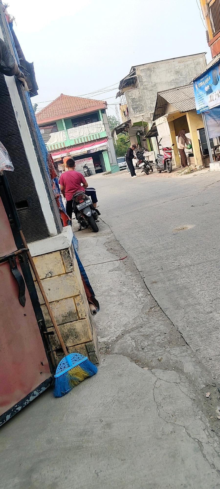 This image depicts a street scene in Mekarmukti, Indonesia. In the foreground, a person in a red shirt is sitting on a black motorcycle. A blue and yellow broom is leaning against a textured stone wall on the left side of the image. The street is paved with asphalt, showing cracks and wear. In the background, there are several buildings, including a two-story building with a reddish-orange tiled roof painted green and white. Another building to the right is painted yellow with a blue awning. Two individuals are visible further down the street; one is standing near a parked motorcycle, and another is standing near a yellow structure, appearing to be engaged in a transaction. The sky is overcast.