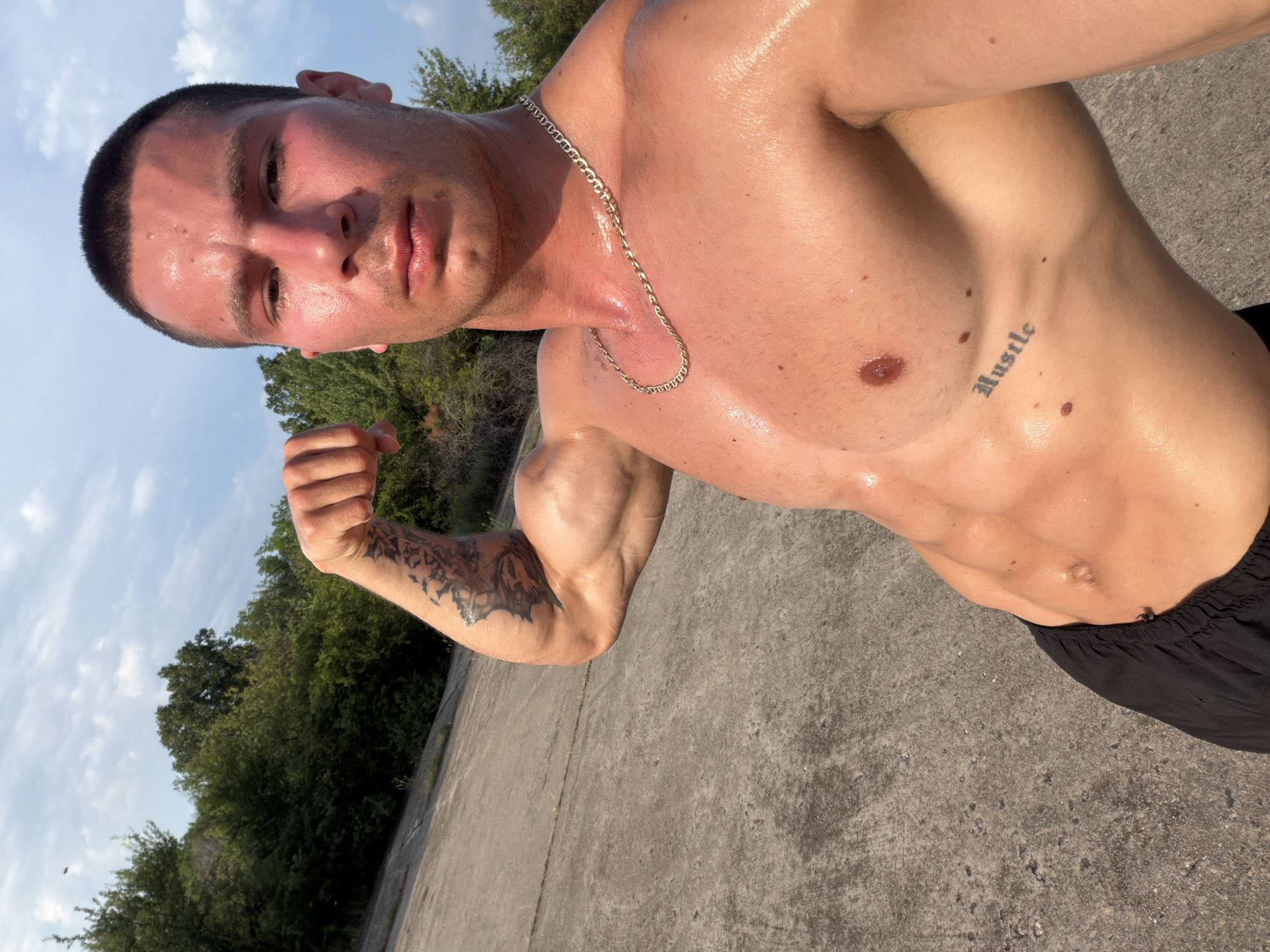 The image features a young adult male captured in a close-up, shirtless selfie outdoors. He exhibits a muscular physique, with visible abdominal definition and a flexed left bicep. His skin appears flushed and glistening with sweat, suggesting recent physical exertion or warm weather. He has short, dark hair and a serious expression. A gold-colored chain necklace with Mariner-style links is worn around his neck. Distinctive tattoos include an intricate pattern on his left forearm/bicep and the word "Hustle" in gothic script on his right ribcage. He is partially clad in black athletic shorts, visible in the lower left quadrant.
The background consists of a sloping, light gray concrete or asphalt surface in the immediate foreground, transitioning to a dense line of green trees and foliage further back. The sky above is light blue with scattered white clouds, indicating daylight and fair weather conditions.
There are no discernible landmarks, signage, or unique architectural or geographical features within the image that allow for the forensic identification of the location as the city of Bogdanitsa, Bulgaria.