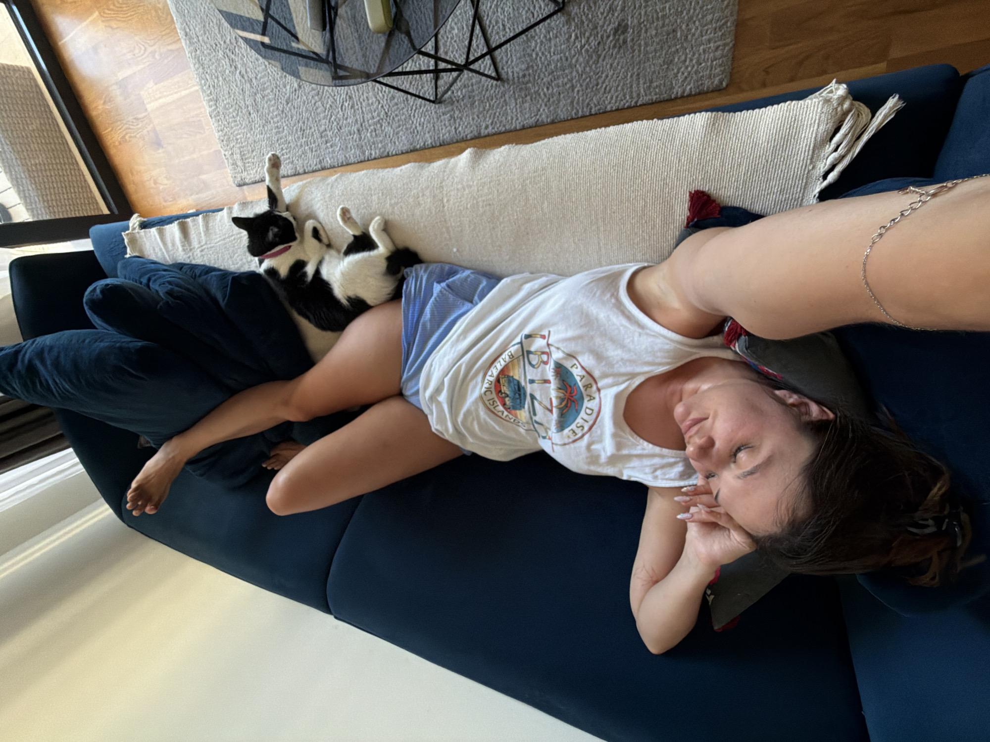 In a sunlit interior, likely an apartment, a woman is lying on a deep blue sofa with her head resting on a pillow. She is wearing a white tank top with a graphic print and blue shorts. Her legs are extended and bent. A black and white cat is lying on the sofa next to her. The sofa is positioned on a light-colored rug. In the background, a large window shows an outdoor balcony with wicker furniture. A modern coffee table with a glass top and metal legs is visible on the rug. The floor is made of polished wood. The scene appears to be in an urban setting, possibly Belgrade, Serbia, based on the architectural style and interior design.