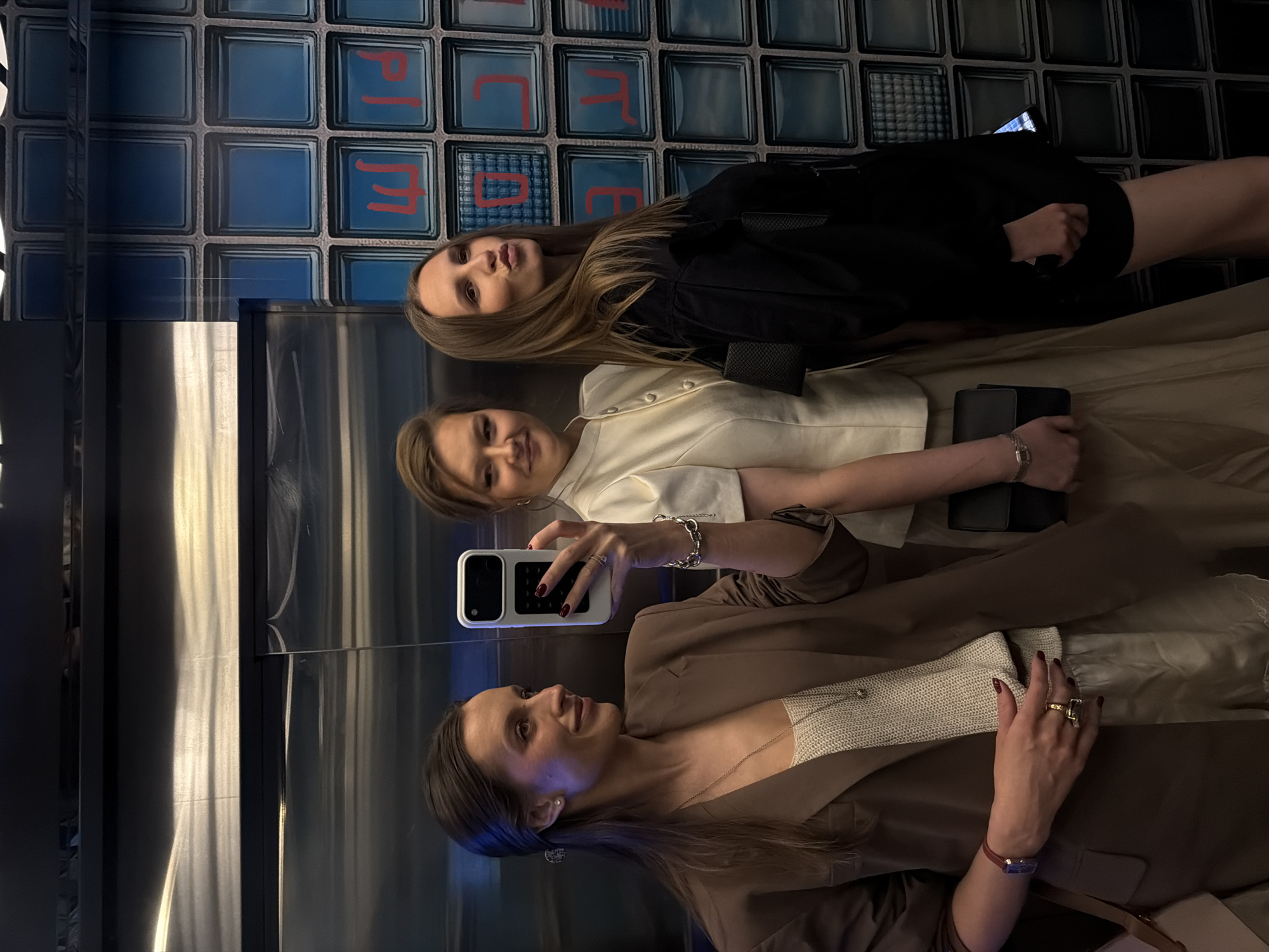 Three women are taking a selfie in an elevator, presumably in Moscow, Russia, given the Cyrillic characters on the background wall. The background features a wall design of blue glass blocks with red Cyrillic letters on some of them. The elevator appears to be paneled with stainless steel. The woman in the foreground wears a brown blazer over a light-colored top and holds a smartphone to take the picture. The second woman wears a white top. The third woman wears a black dress. All are looking at the camera.